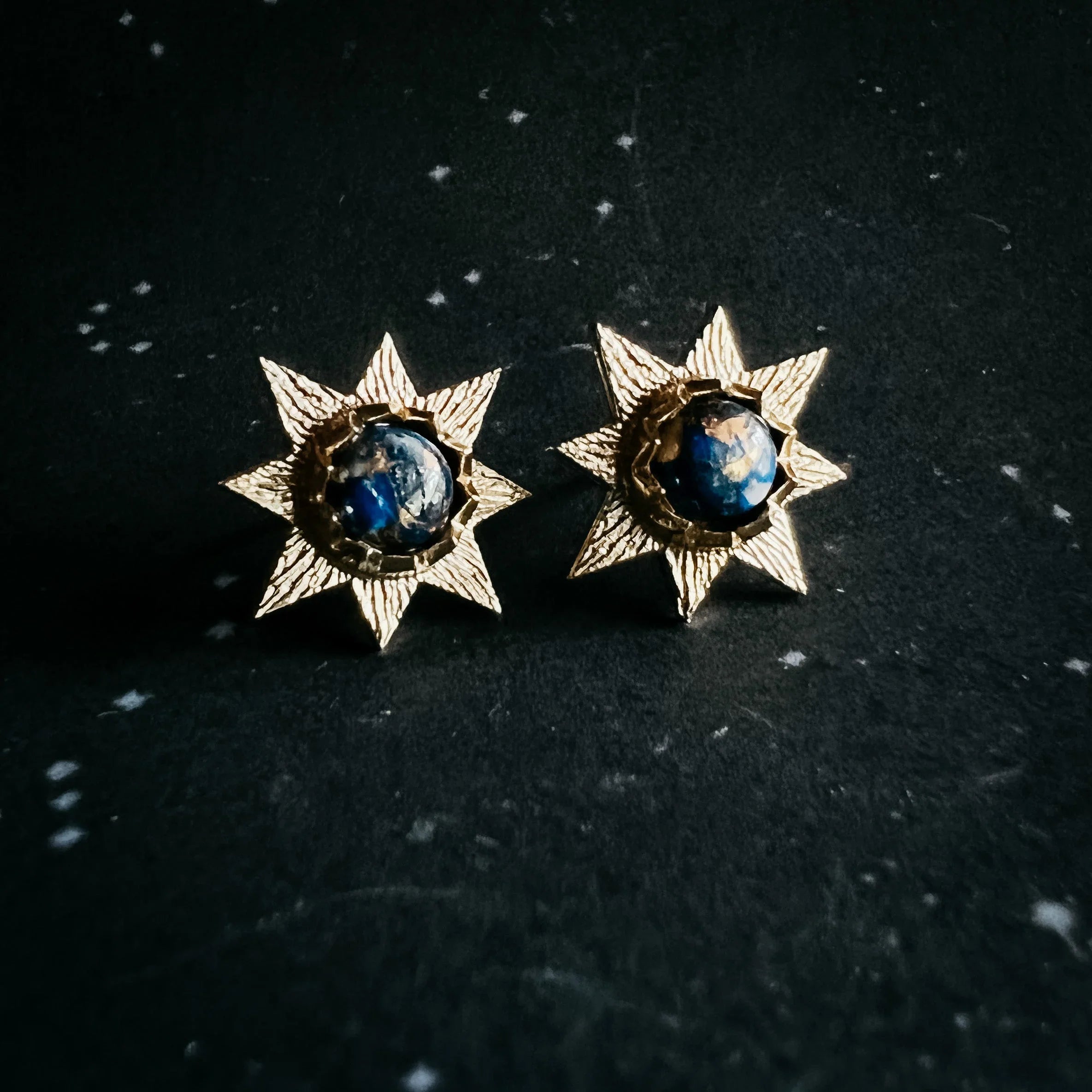 Gold starburst earrings with cosmic blue centers on a black speckled background at Qu!rk Boutique
