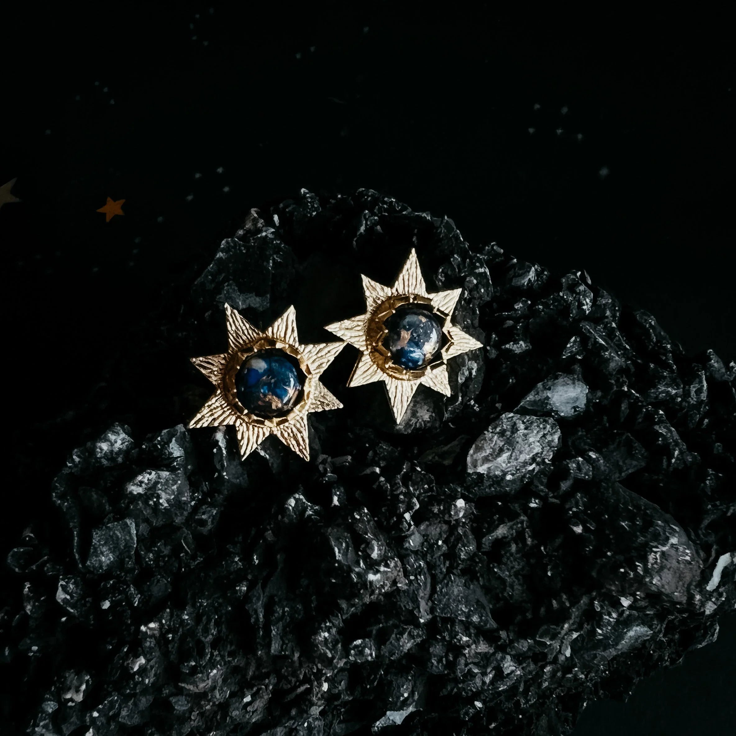 Gold starburst earrings with cosmic blue centers on black textured stone, Qu!rk Boutique