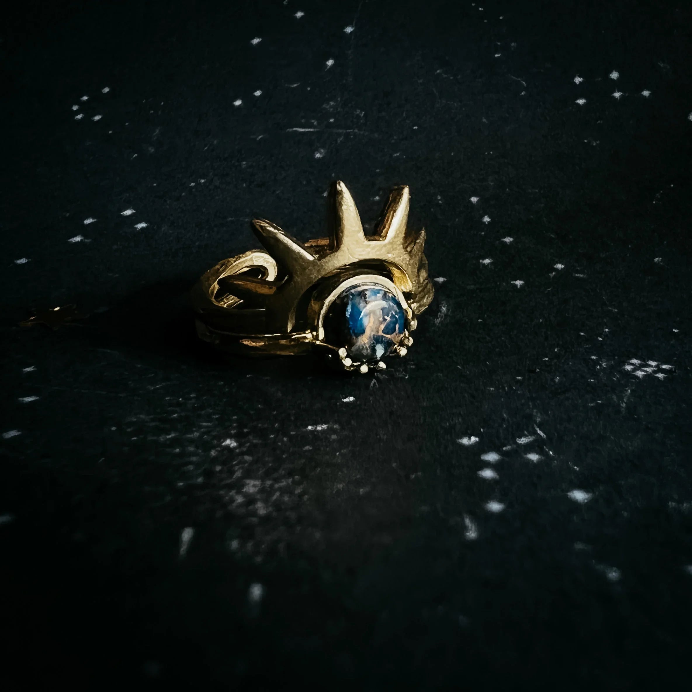 Gold ring with blue gemstone and cosmic crown design on a star-speckled black surface