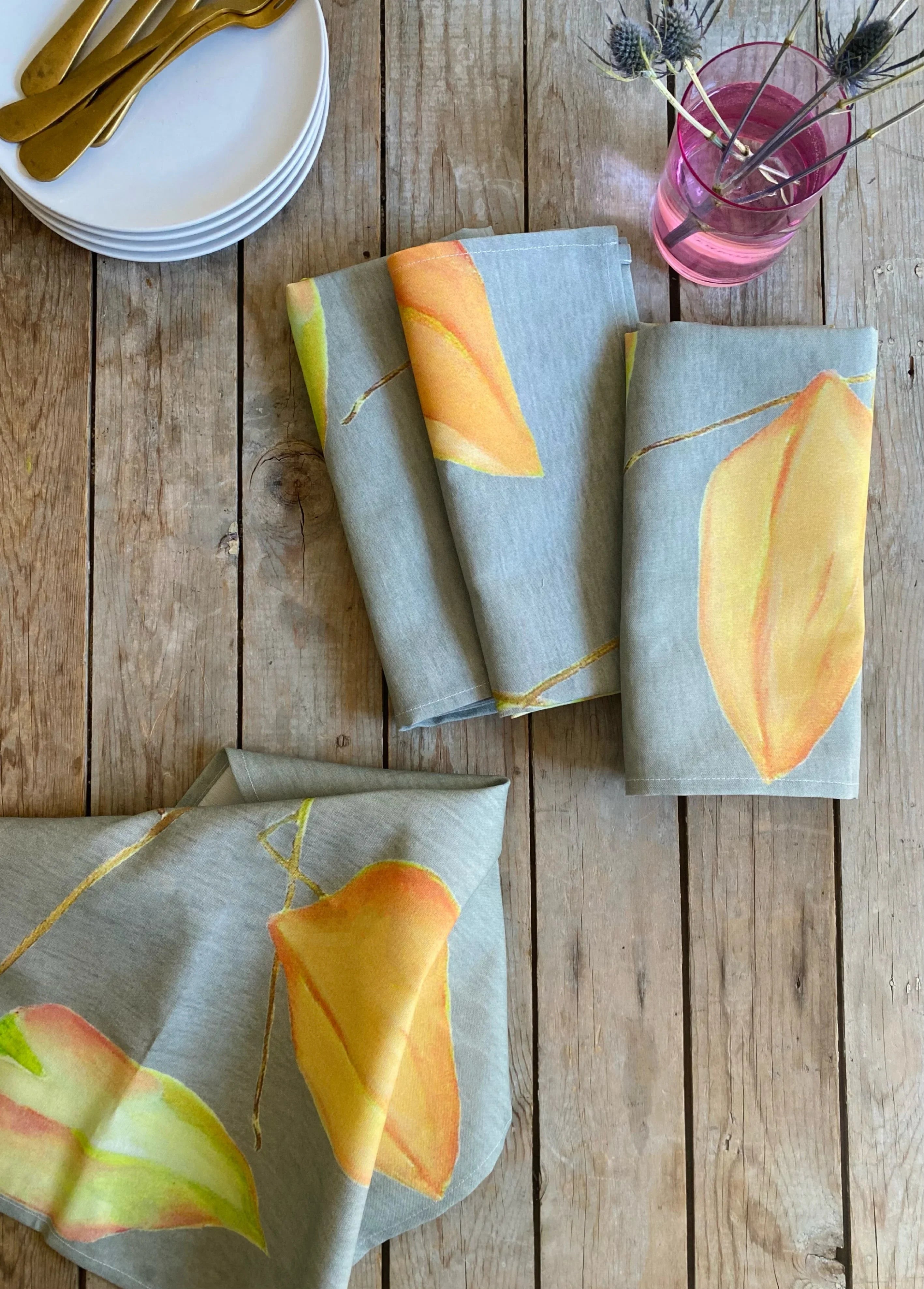 Folded gray cotton napkins with orange-yellow floral print on rustic wooden table with white plates and golden cutlery