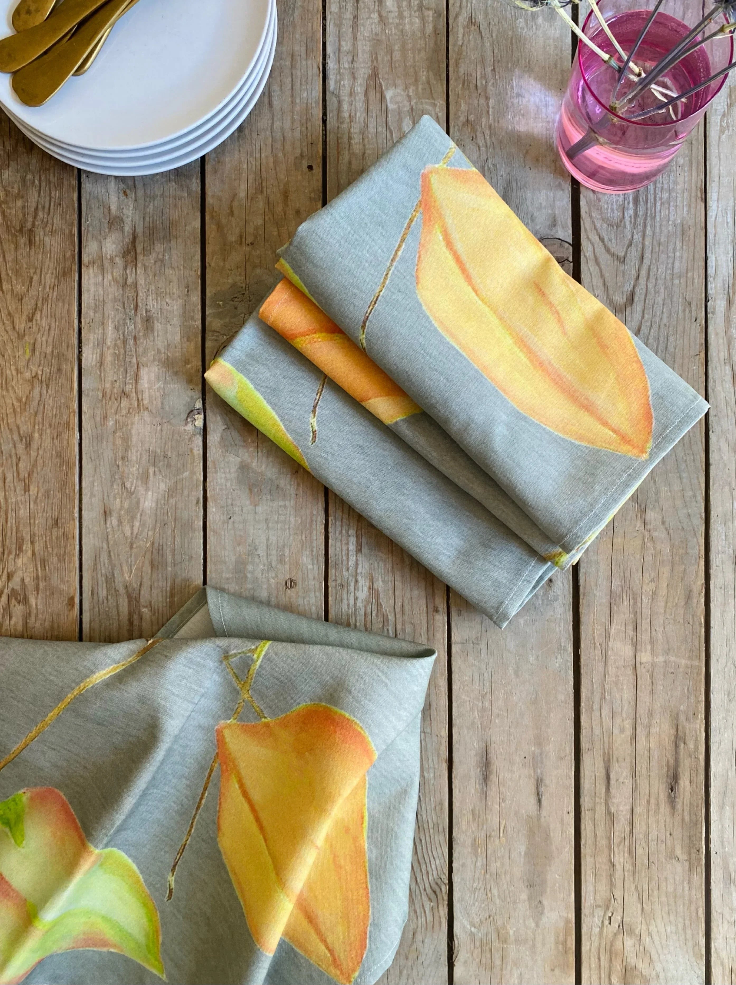Folded gray tablecloth with yellow and orange leaf patterns on rustic wooden table with white plates and pink glass vase