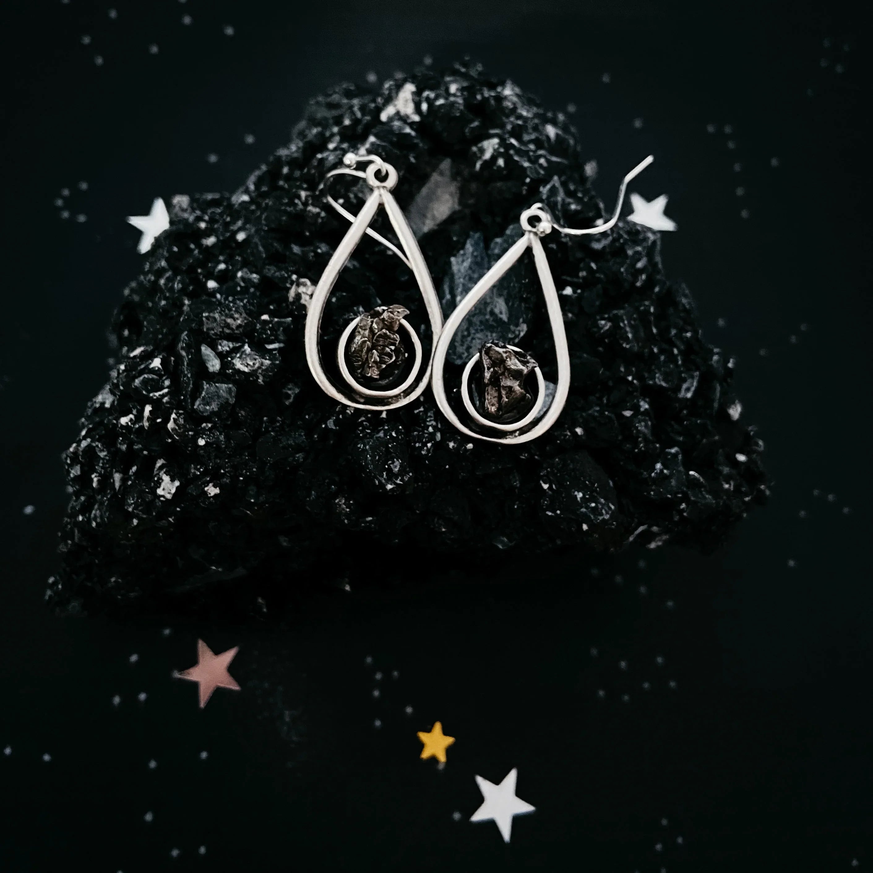 Silver teardrop earrings with dark textured stones displayed on black volcanic rock with star-shaped confetti