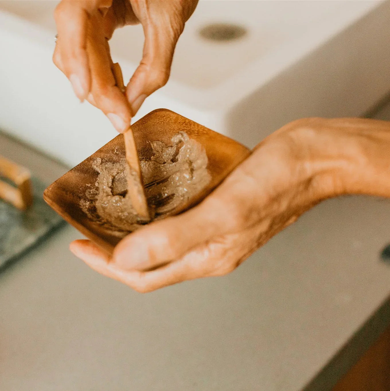 Hands mixing a scrub or mask in a small wooden bowl with a wooden spatula on bathroom counter