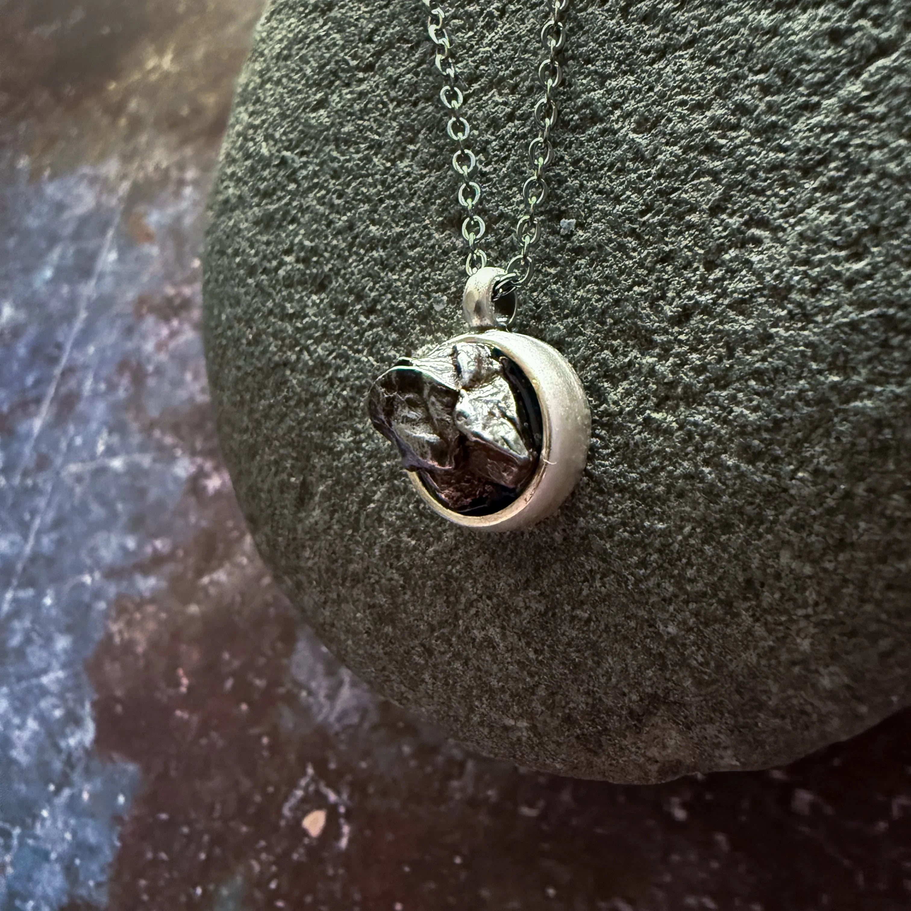 Silver chain necklace with a round pendant featuring an irregular metallic stone on a textured grey rock