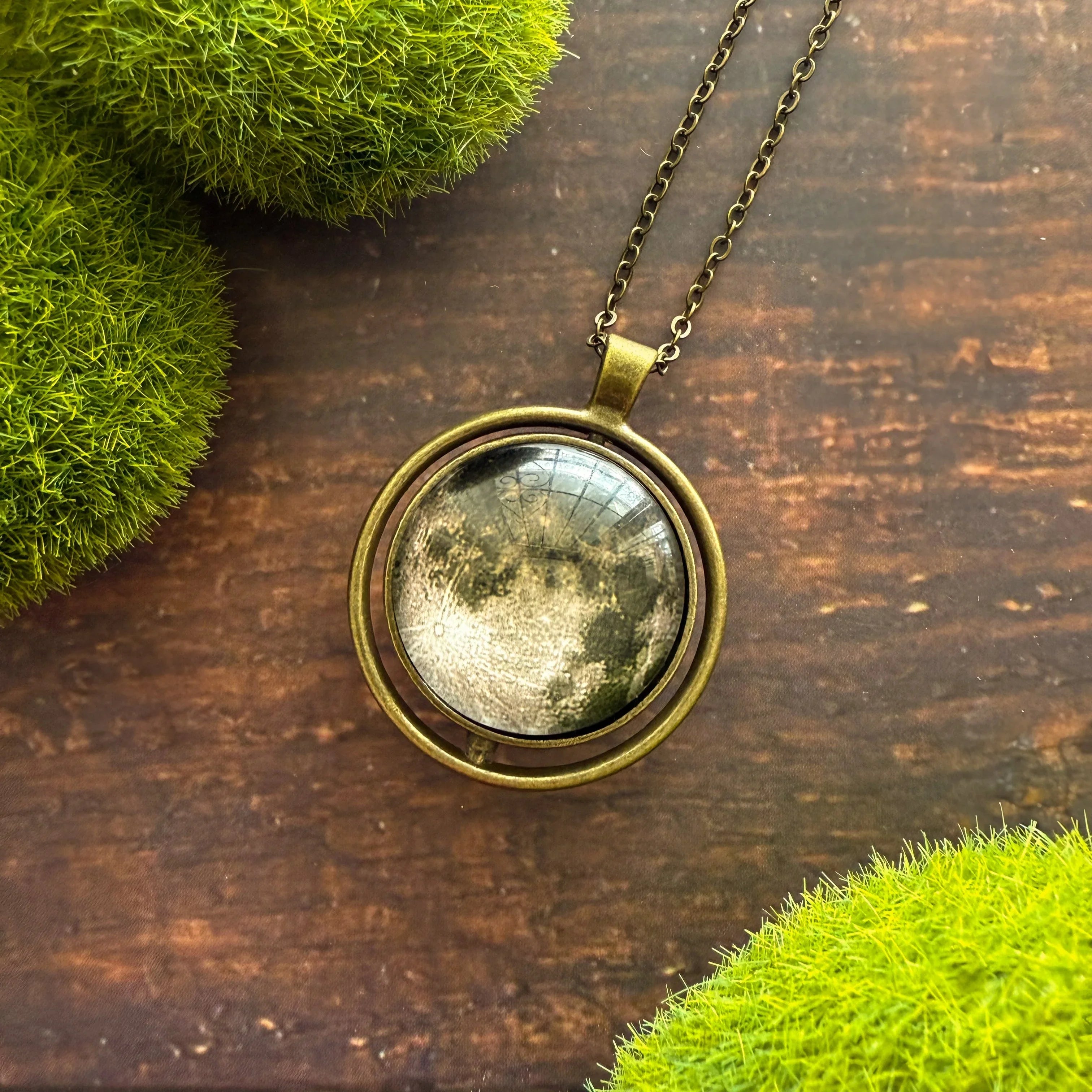 Moon phase pendant necklace on wood, surrounded by green moss, cosmic boutique jewelry