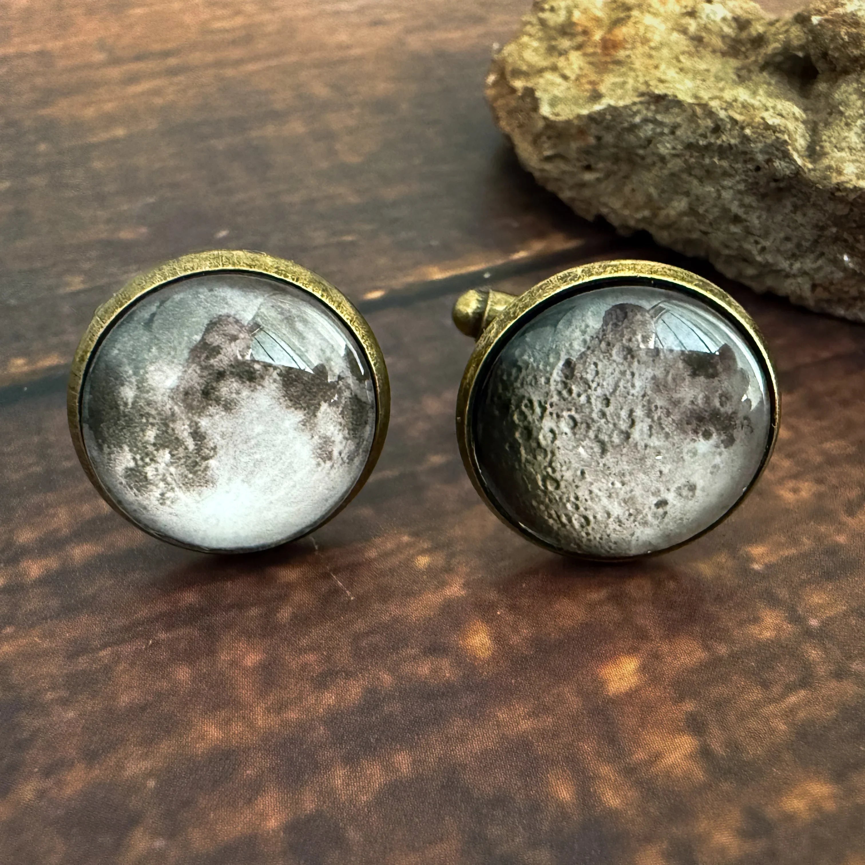 Round moon-themed cufflinks with brass finish placed on rustic wooden surface near a textured rock