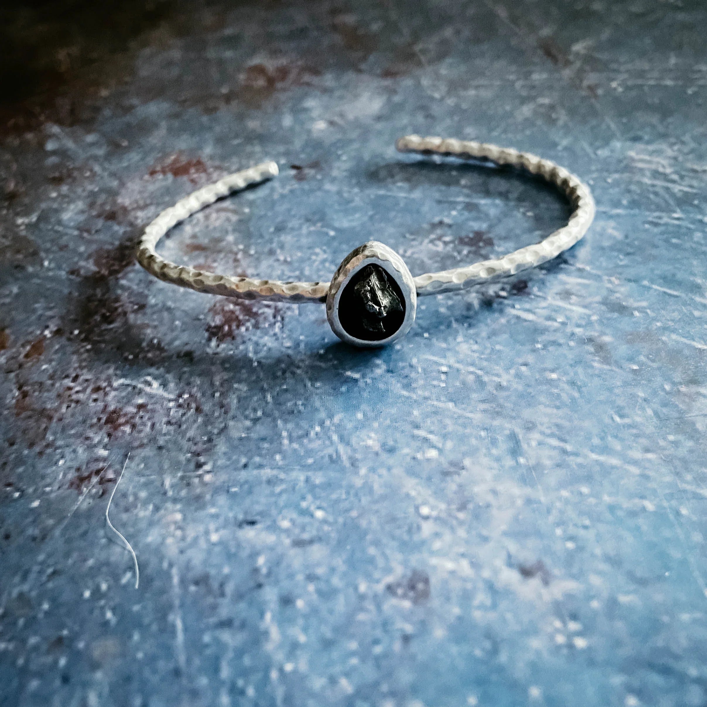 Hammered silver cuff bracelet with black teardrop stone on textured blue surface