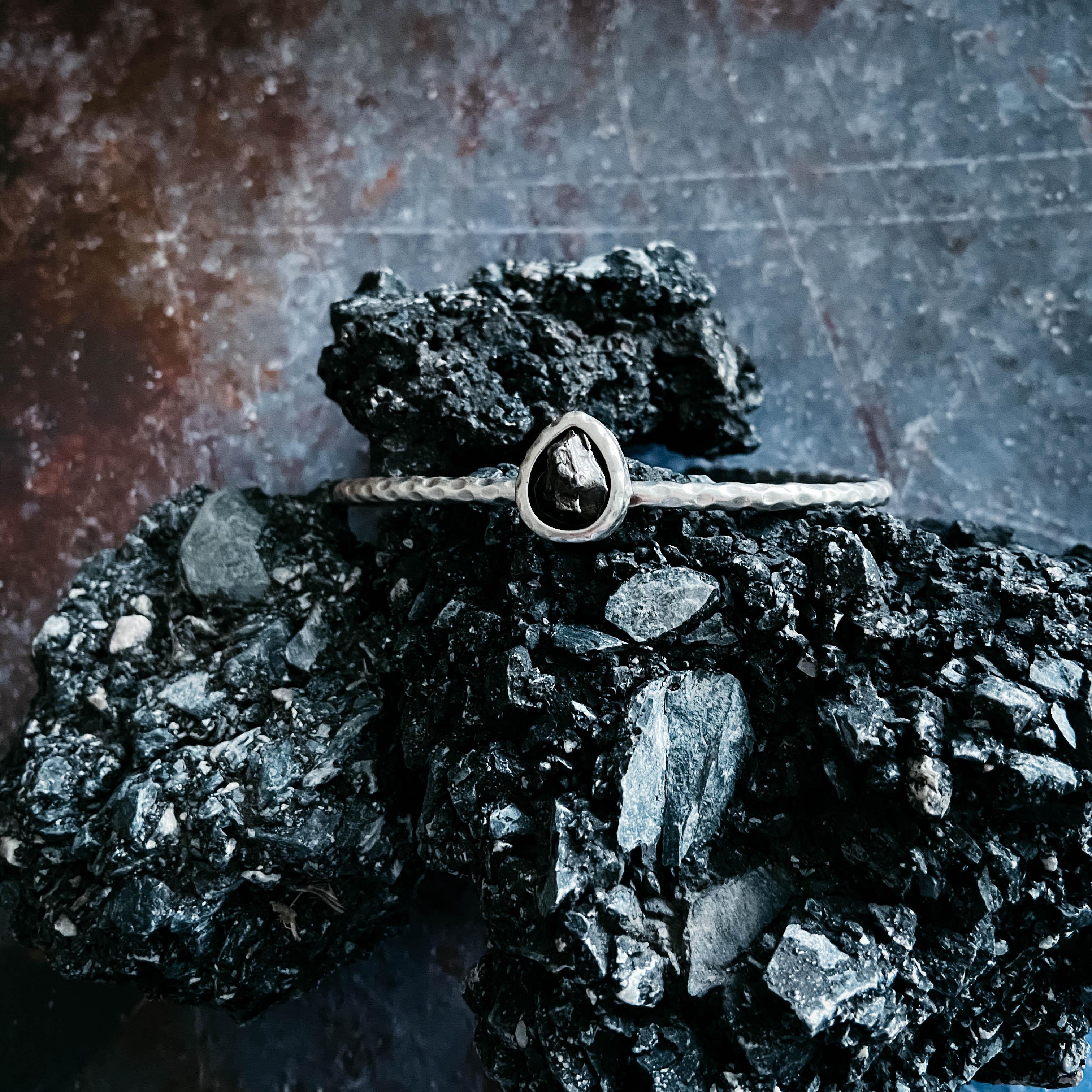 Hammered silver bracelet featuring a rough black stone, displayed on textured black volcanic rocks