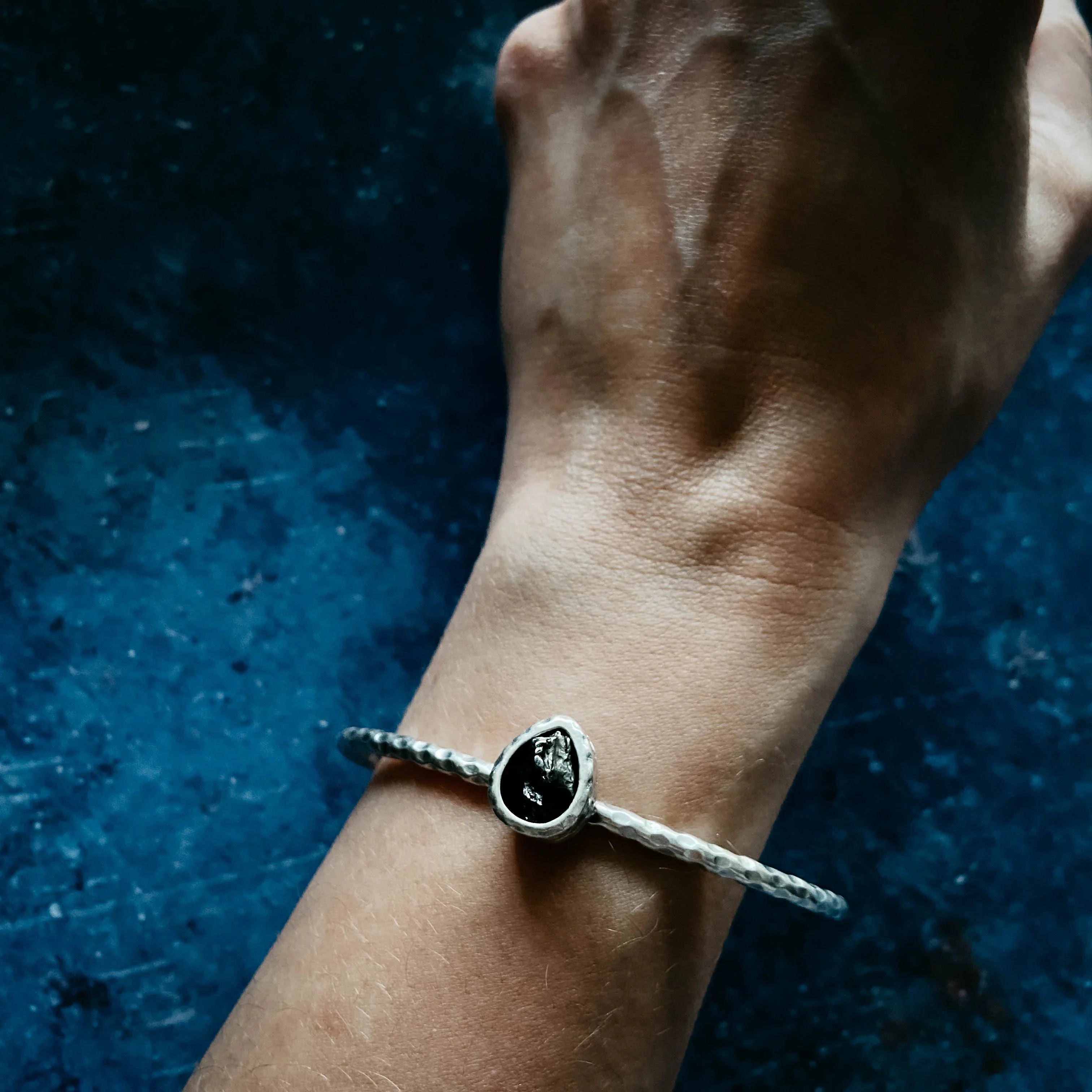 Hand wearing hammered silver bangle bracelet with black stone on textured dark blue background