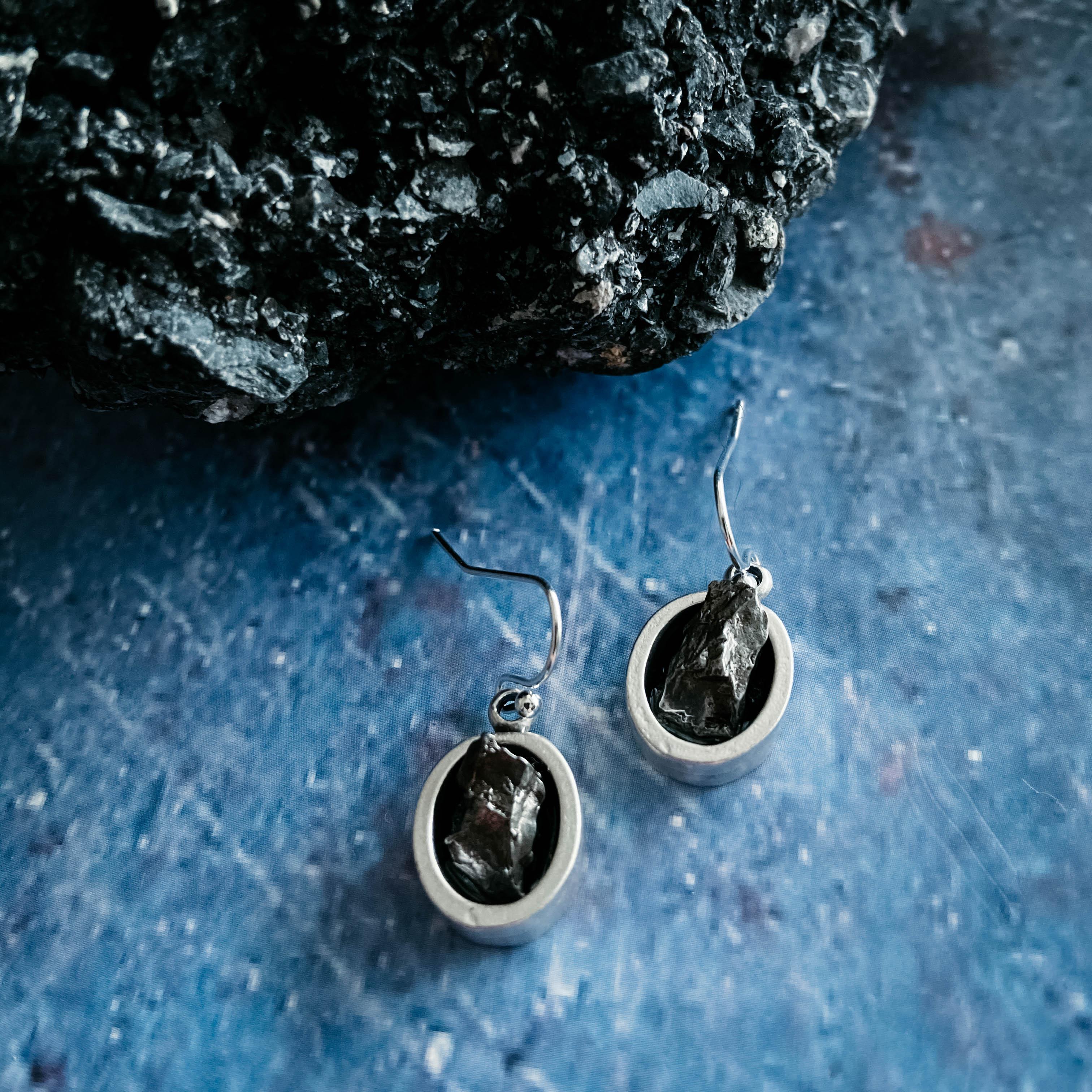 Raw stone dangle earrings with silver oval frames on blue surface, cosmic boutique style.