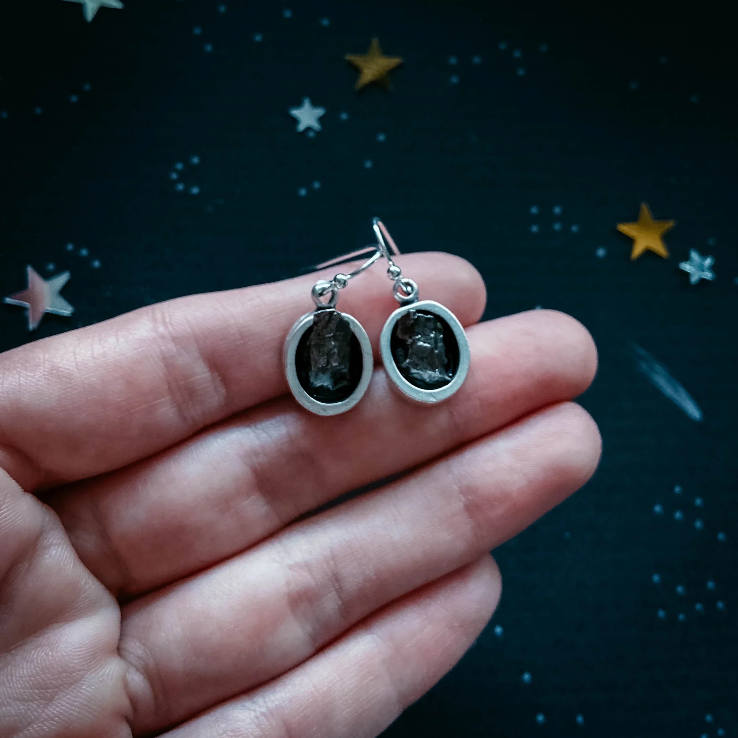 Hand holding cosmic-inspired black stone dangle earrings, starry background, Qu!rk Boutique