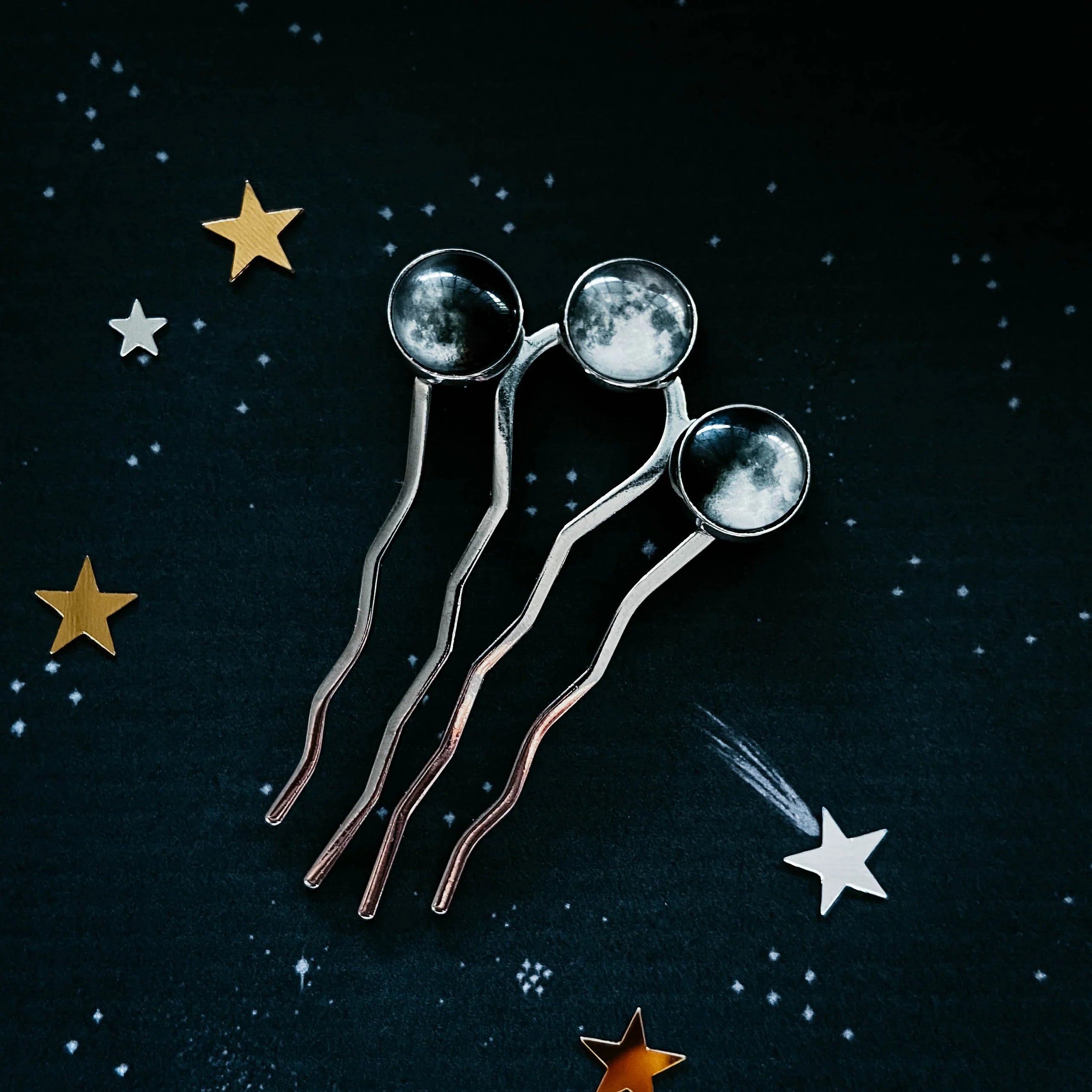 Three silver hairpins with round moon phases on dark starry background and golden star decorations