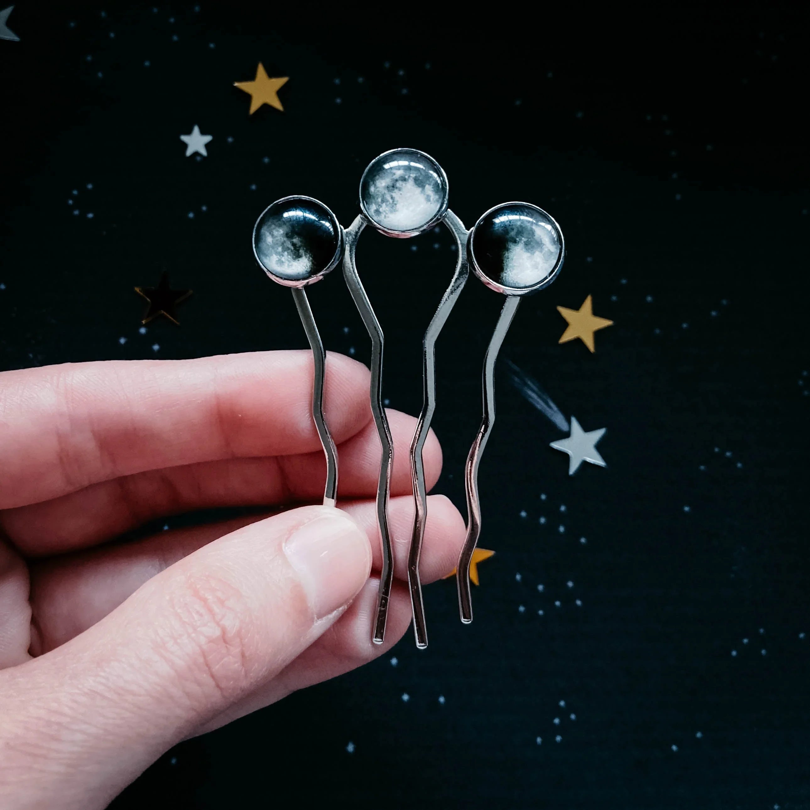Hand holding three silver hairpins with moon phase glass cabochons on a starry background