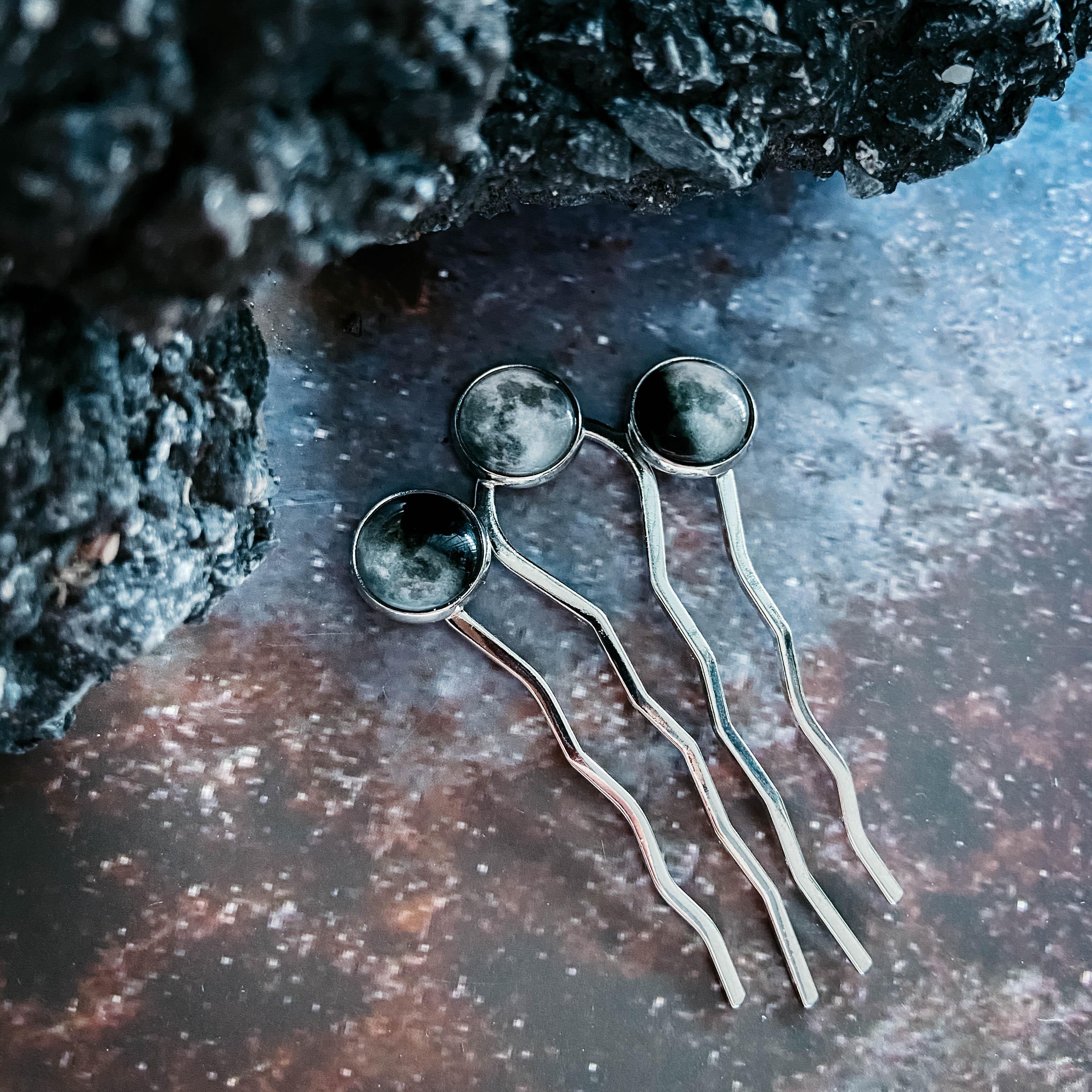 Set of three silver hairpins with moon phase designs on a textured dark surface next to black lava rock