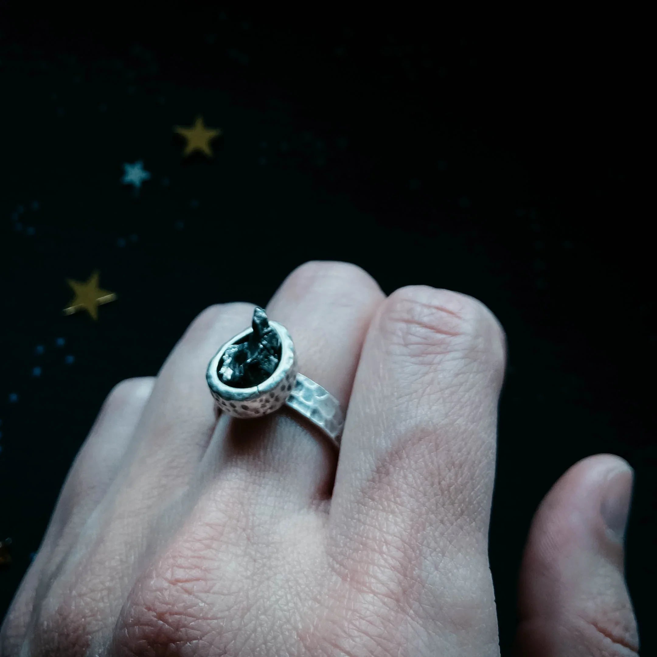 Hand wearing a hammered silver ring with a dark stone, cosmic star background, Qu!rk Boutique