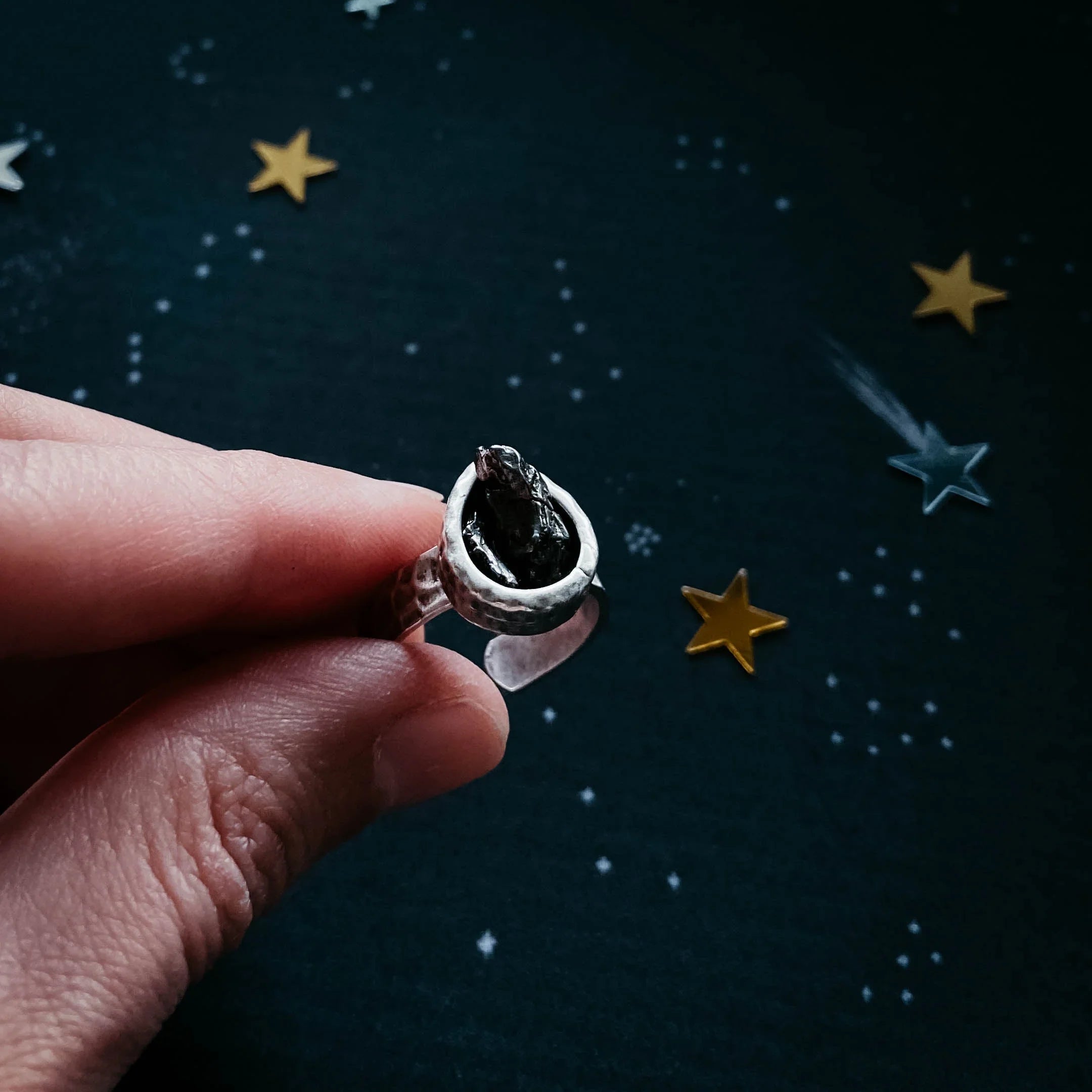 Hand holding cosmic-inspired artisan ring with black stone, star confetti background, Qu!rk Boutique
