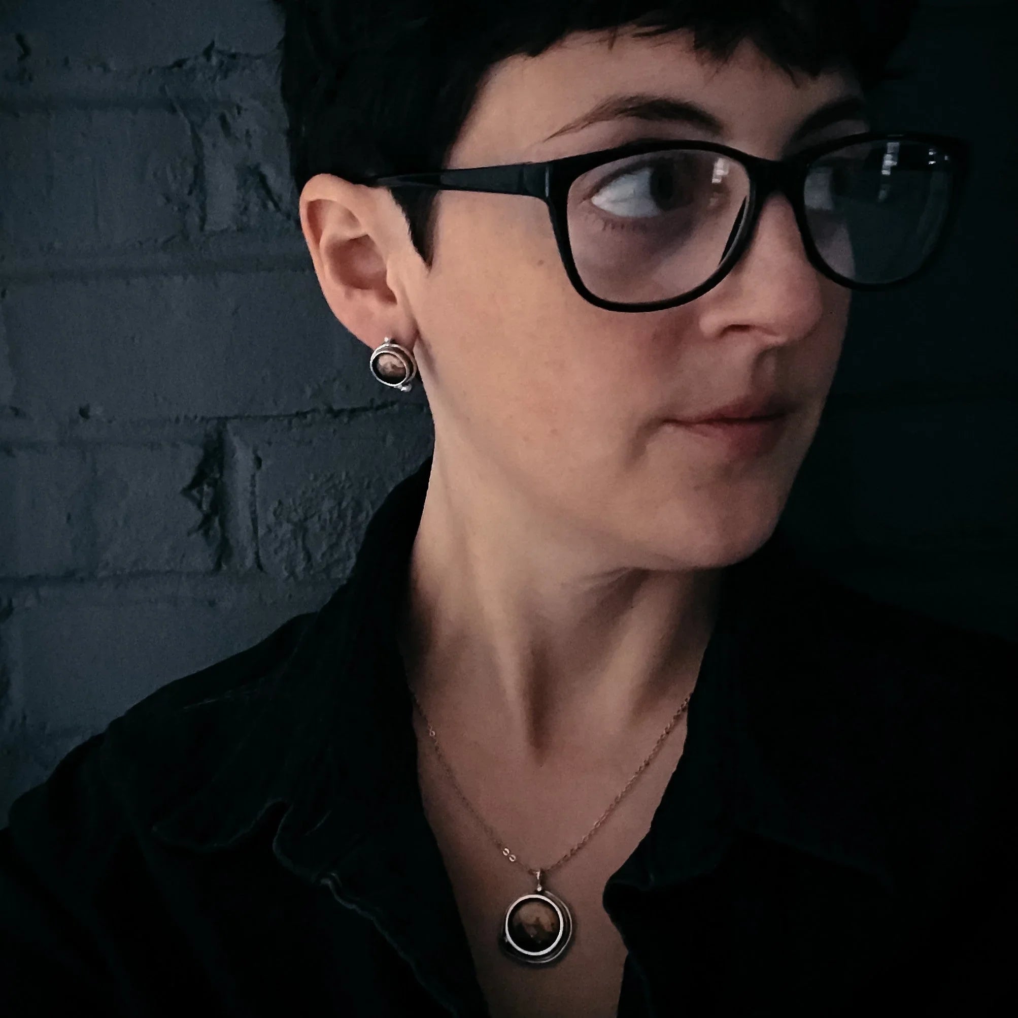 Person with short dark hair wearing black glasses, silver earrings, and pendant necklace against dark brick wall