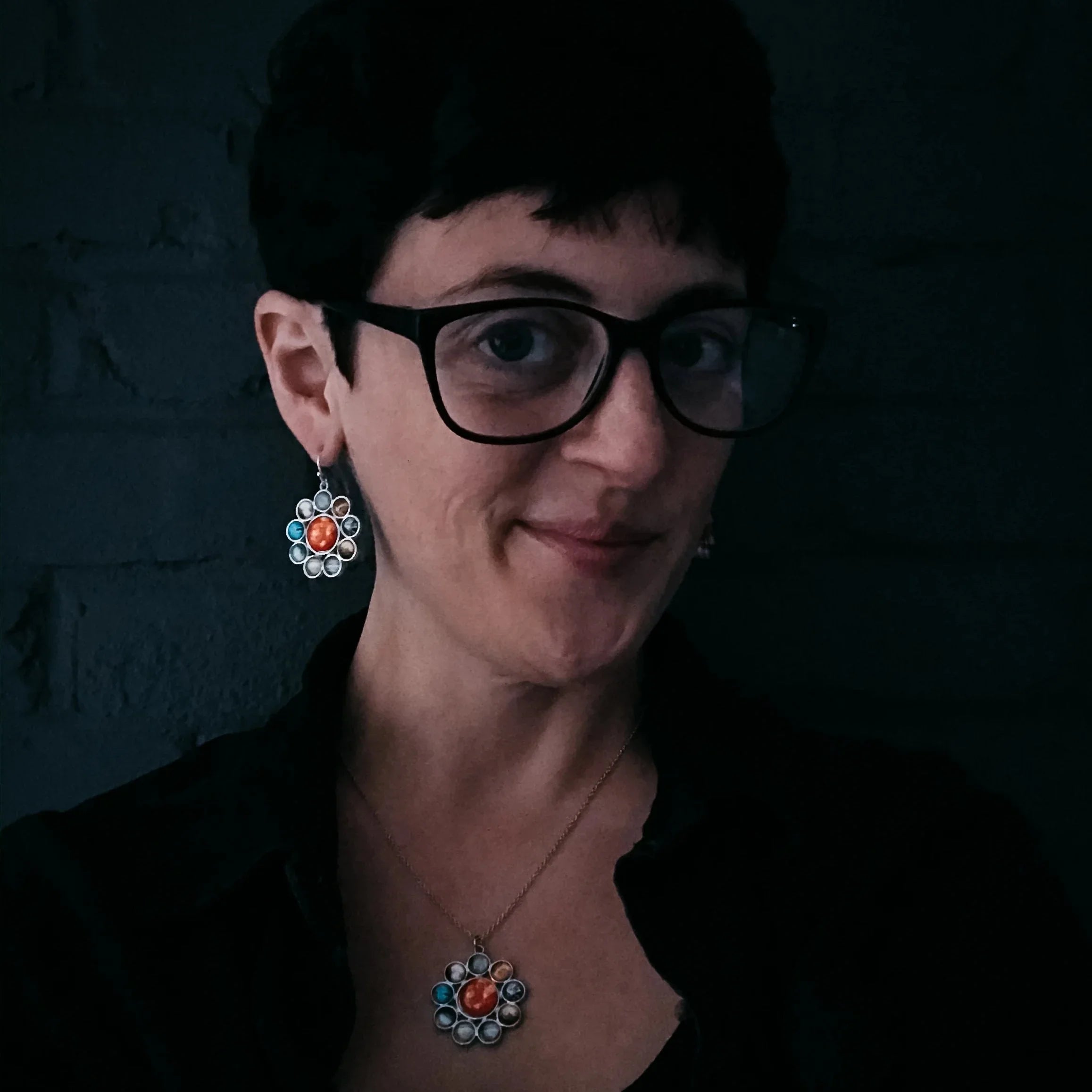 Woman with short dark hair, black glasses, wearing matching multicolored stone earrings and pendant necklace in dark setting