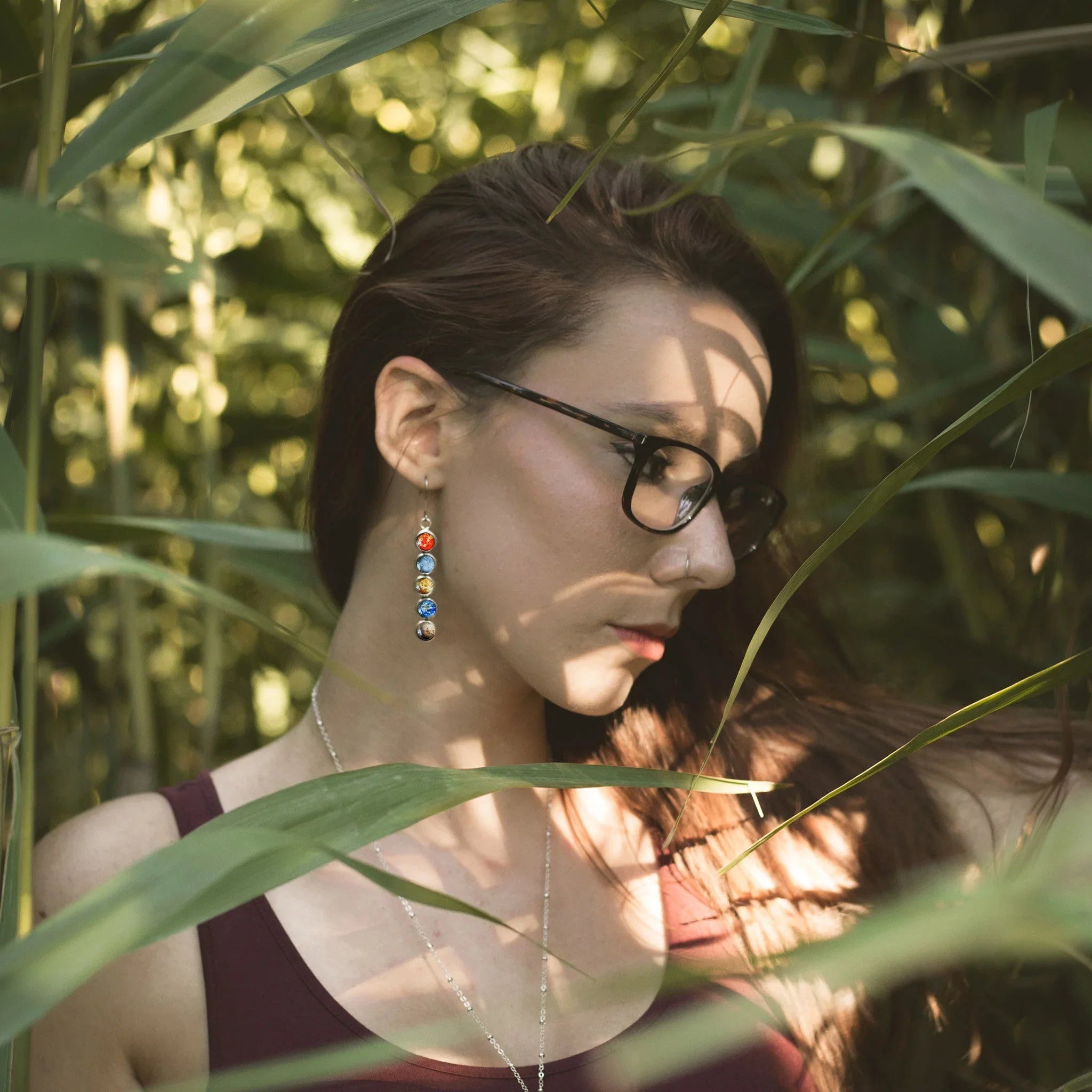 Woman with glasses and colorful planet earrings in nature surrounded by green leaves and light shadows