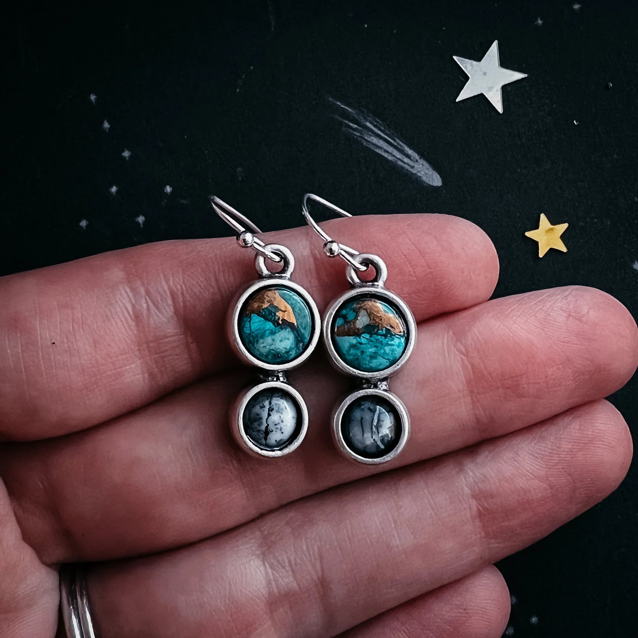 Hand holding silver dangle earrings with turquoise and gray round stones on black starry background