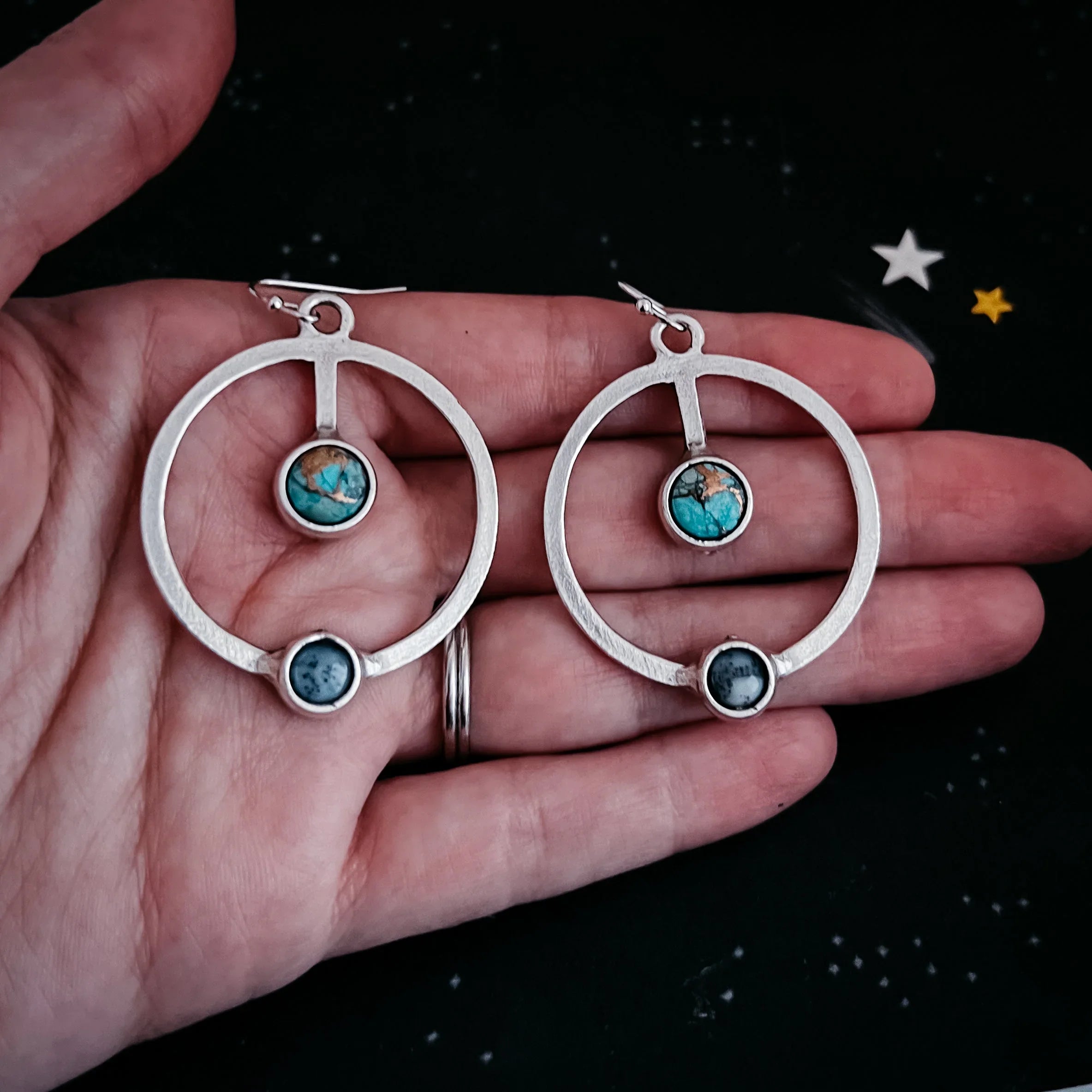 Hand holding silver hoop earrings with turquoise and dark round stones on black starry background