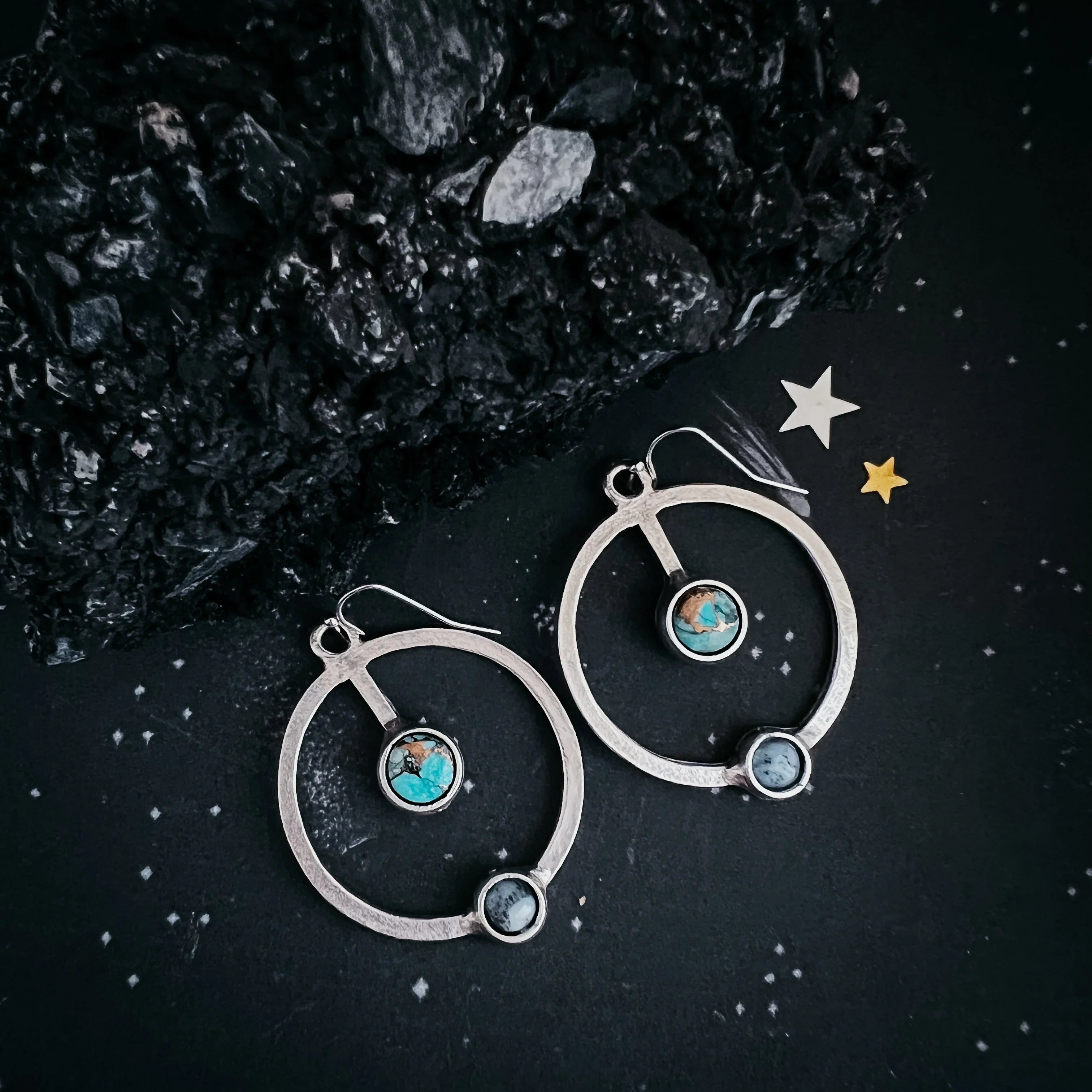 Silver hoop earrings with turquoise and gray stone accents on a dark textured surface with star decorations