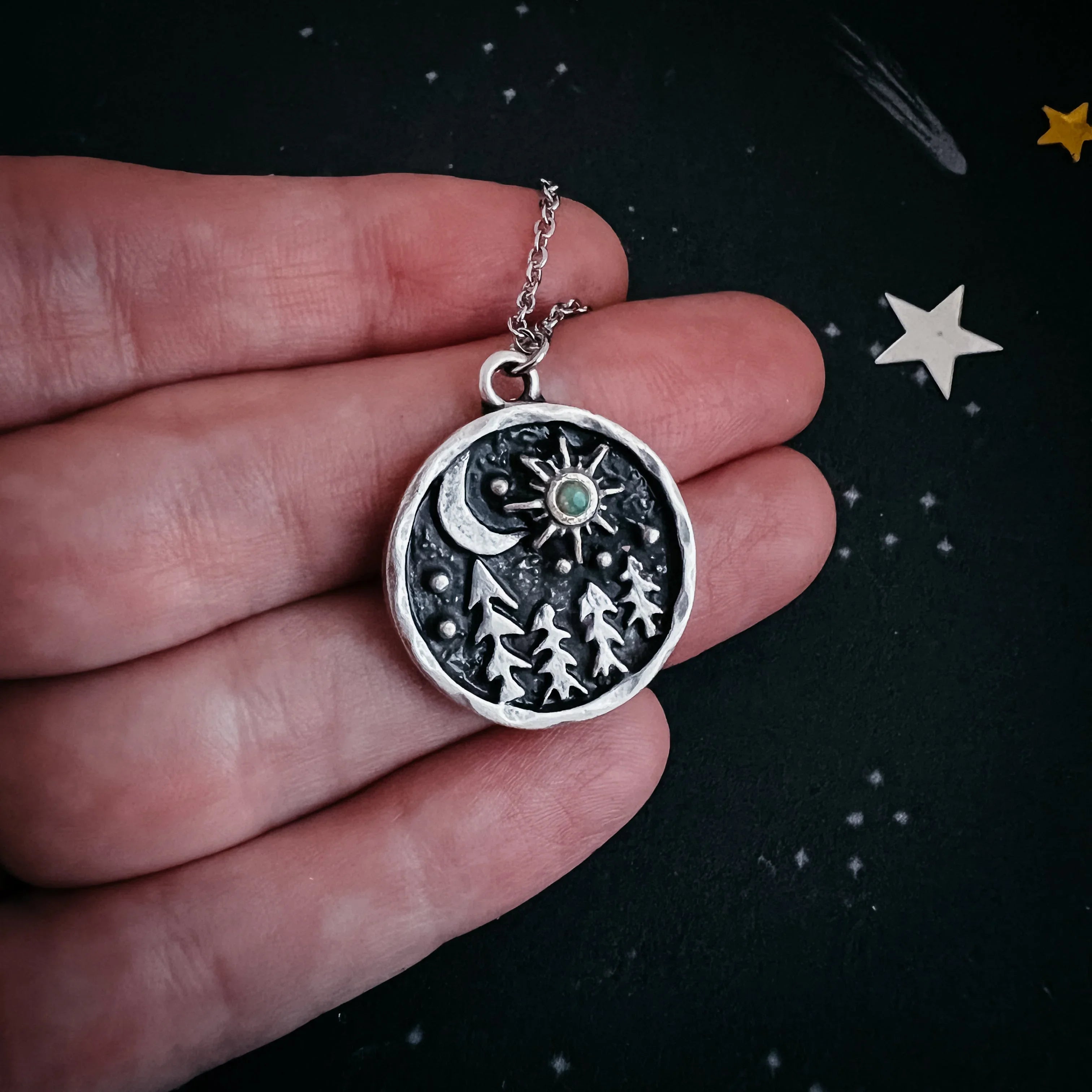 Hand holding artist-designed silver pendant necklace with forest, moon, and star details