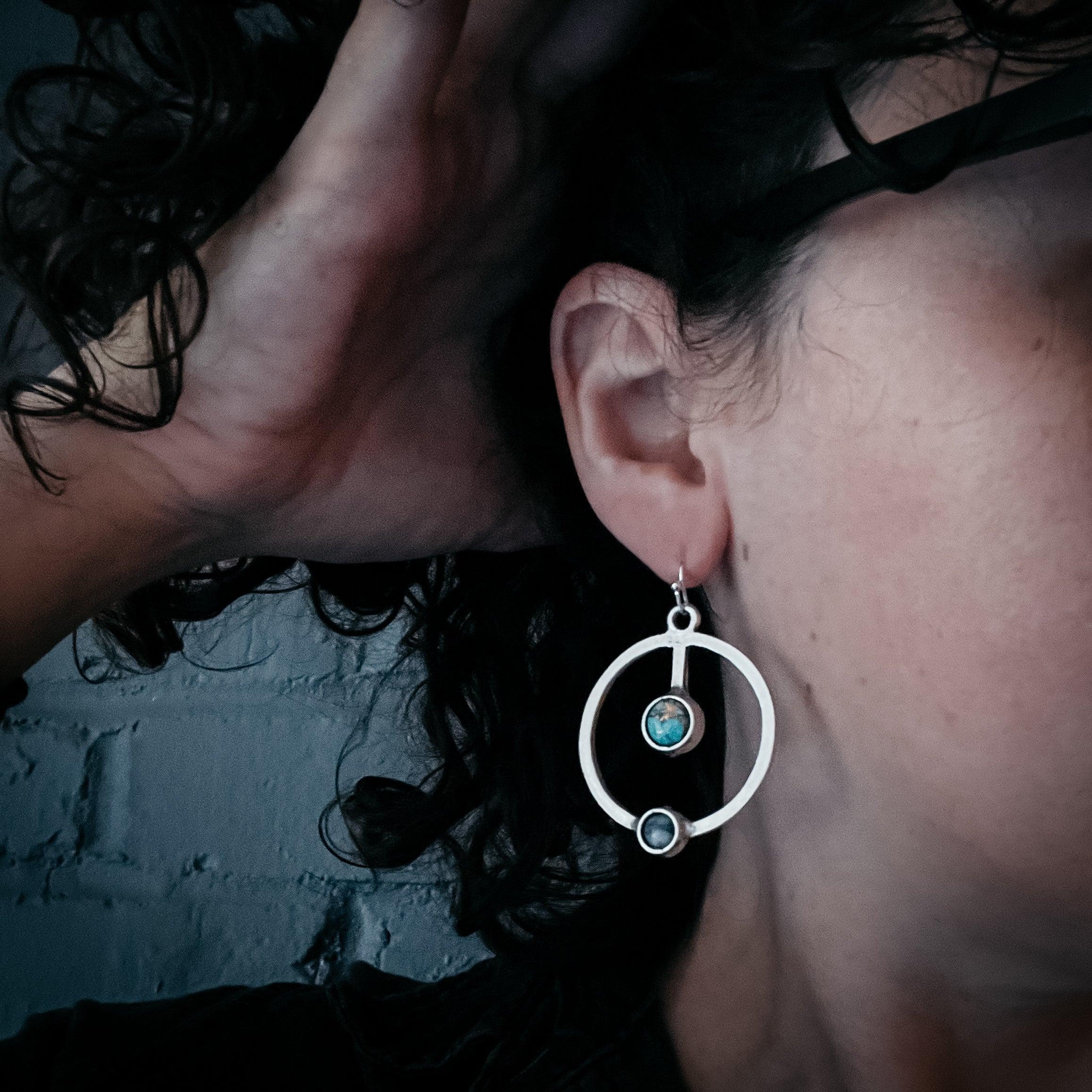 Close-up of person wearing silver hoop earrings with blue stones against dark curly hair and grey brick wall