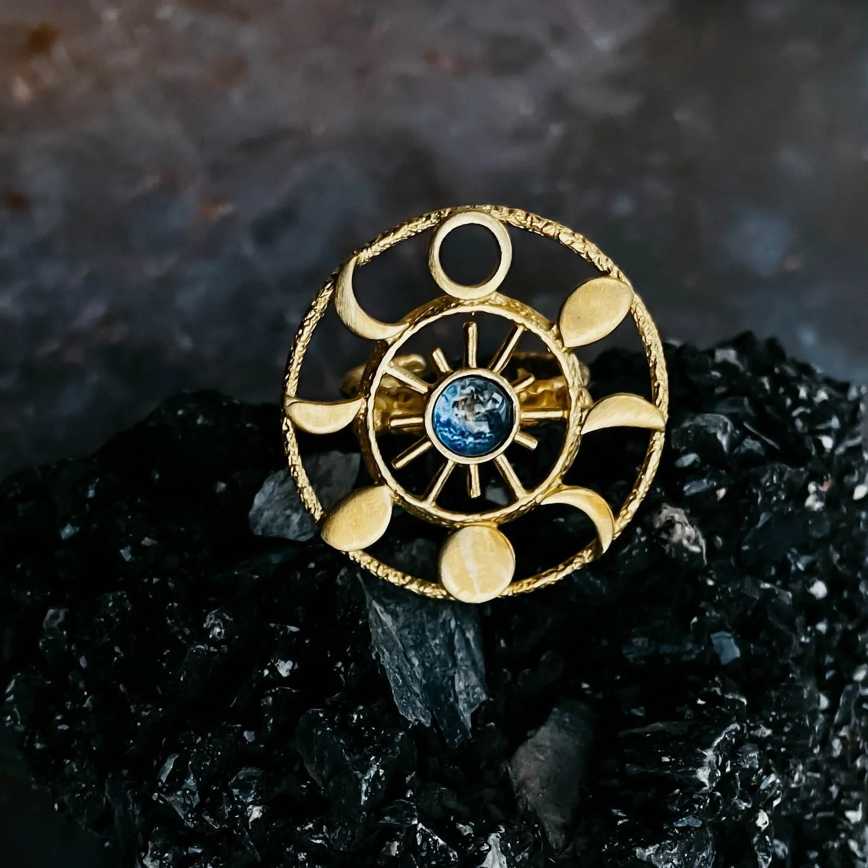 Gold ring with sun and moon phases design, featuring a blue gemstone center on black textured surface