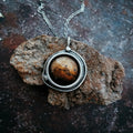 Round brown gemstone pendant in silver setting on chain, resting on textured brown rock