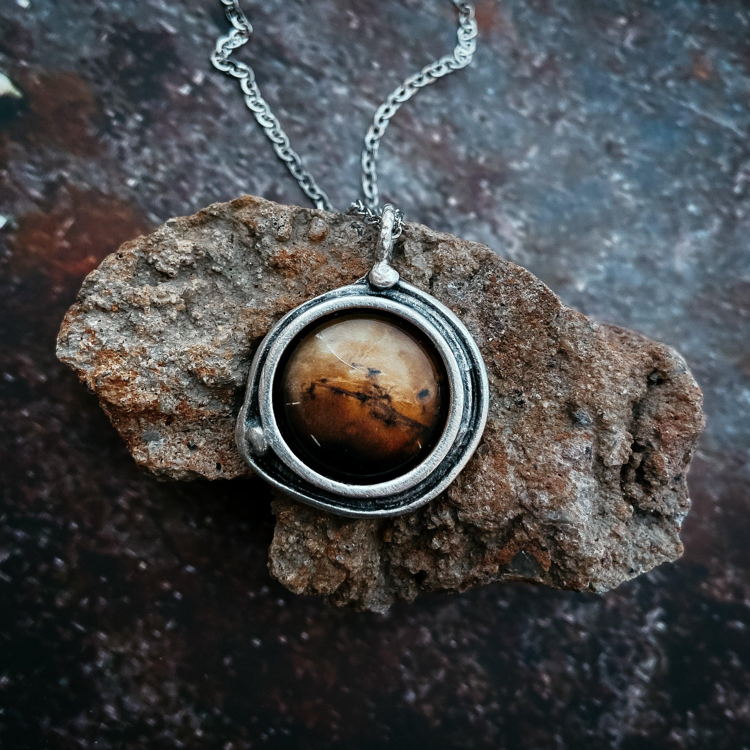 Round brown gemstone pendant in silver setting on chain, resting on textured brown rock