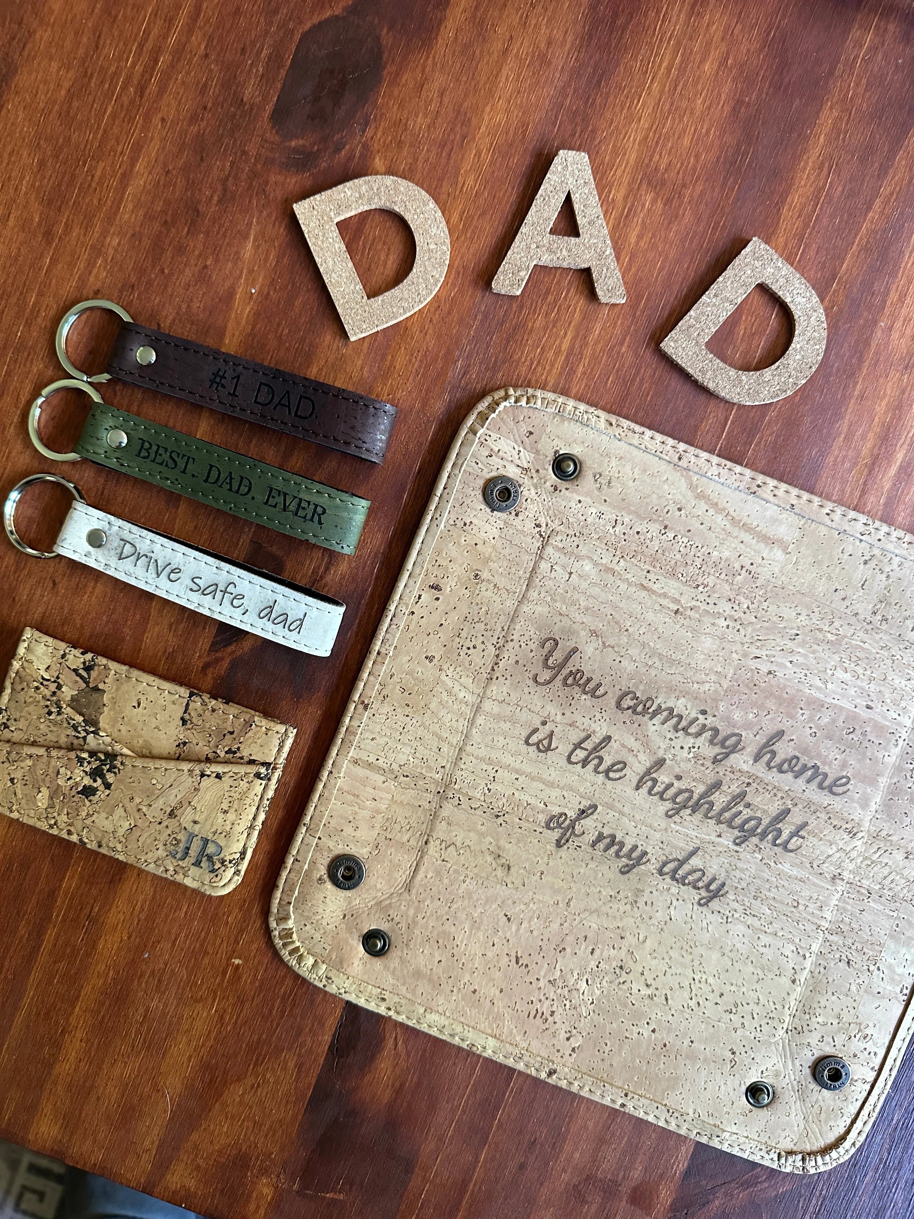 Eco-friendly dad gifts on wood: cork wallet, engraved keychains, and 'You coming home' pouch.