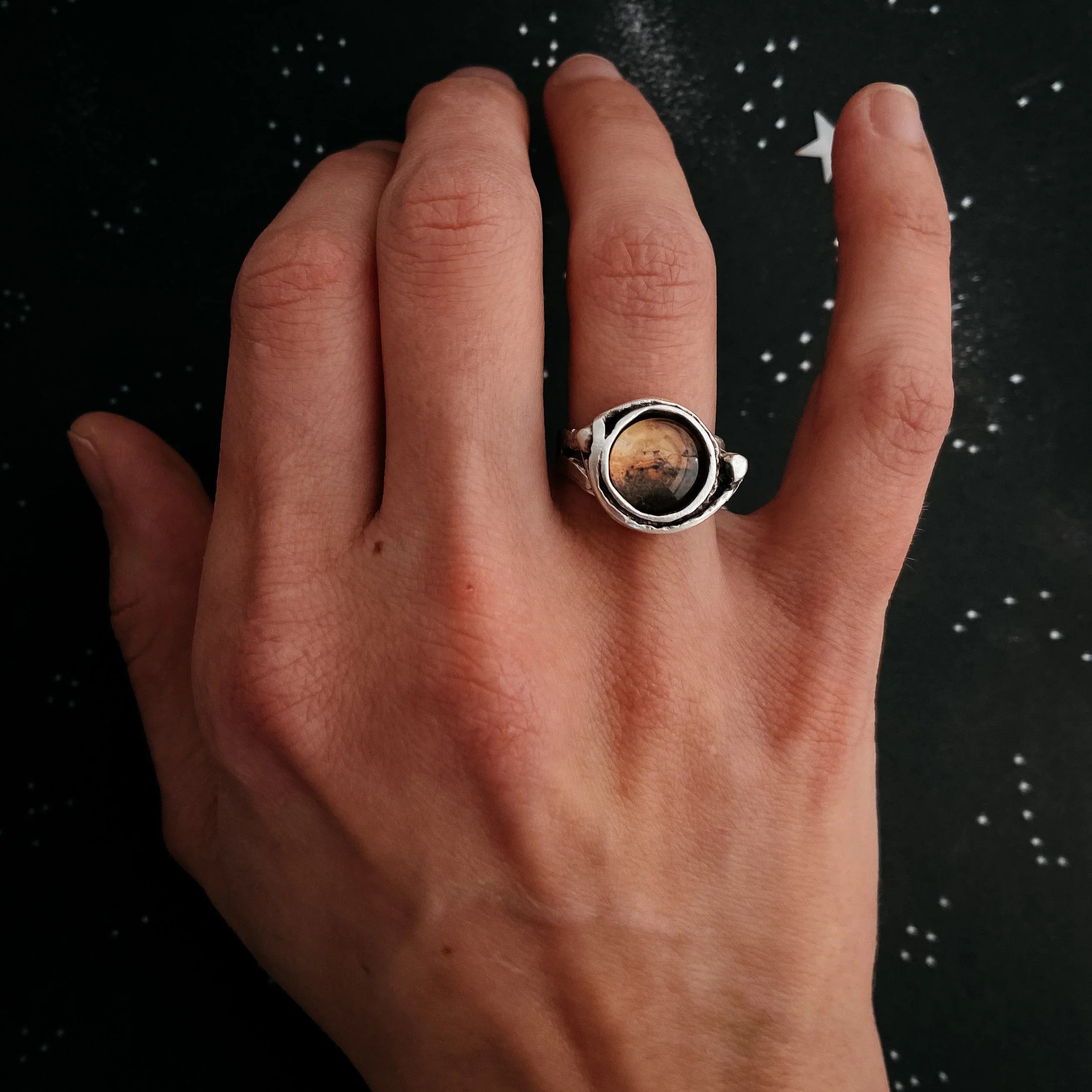 Hand wearing a silver ring with a round, amber-colored stone on a starry black background