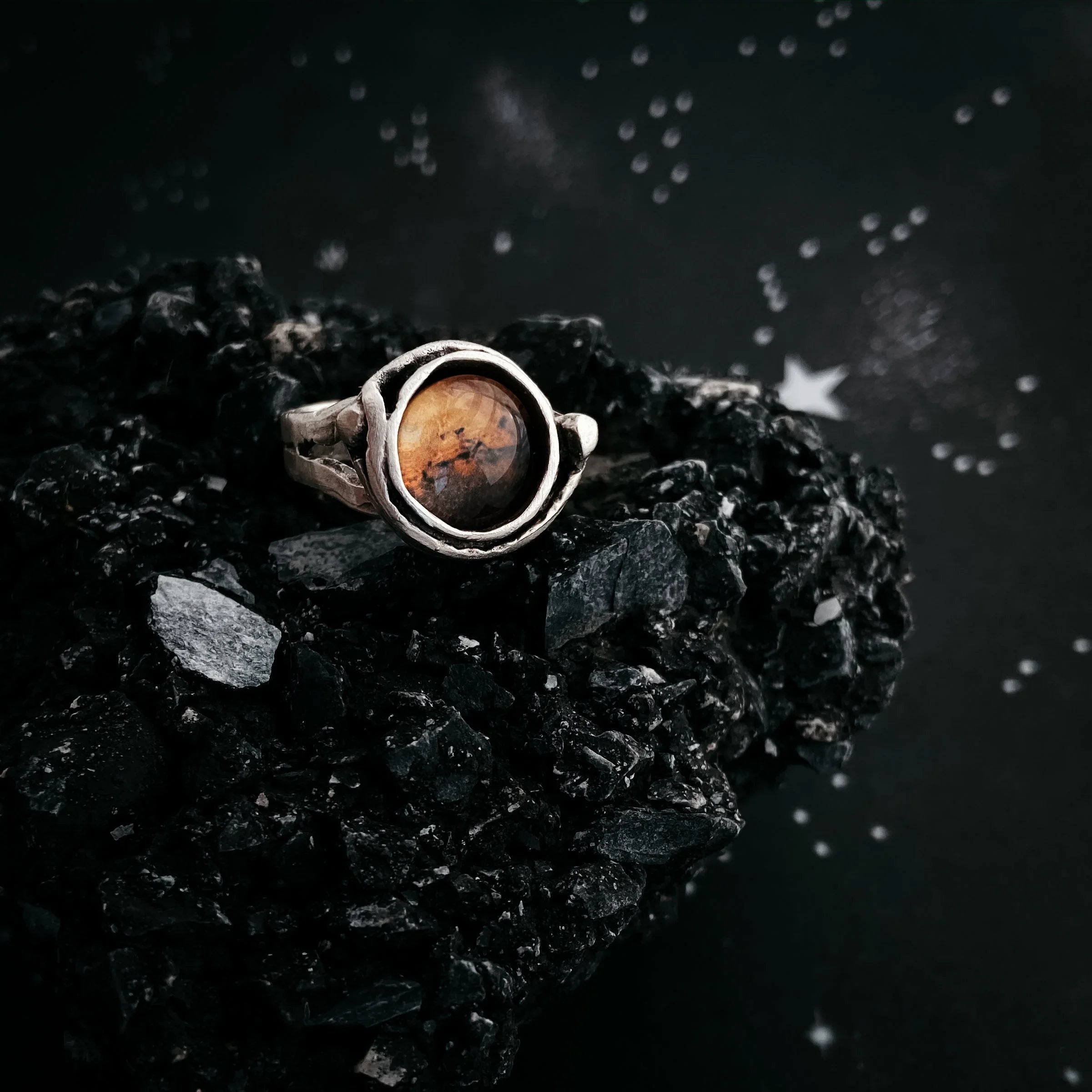 Silver ring with round amber stone on black textured rock with starry background