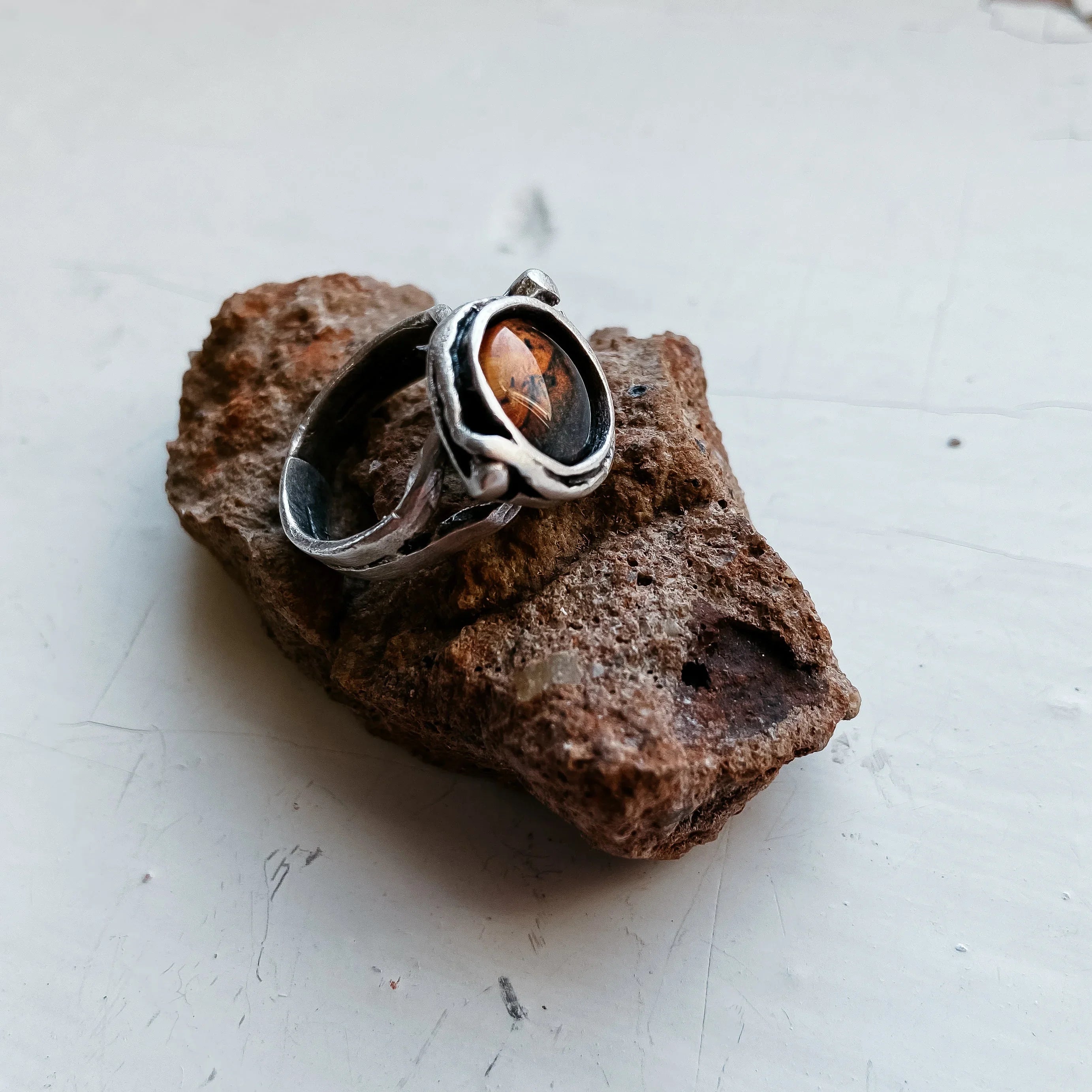 Silver ring with brown gemstone resting on a rough textured brown rock on white surface
