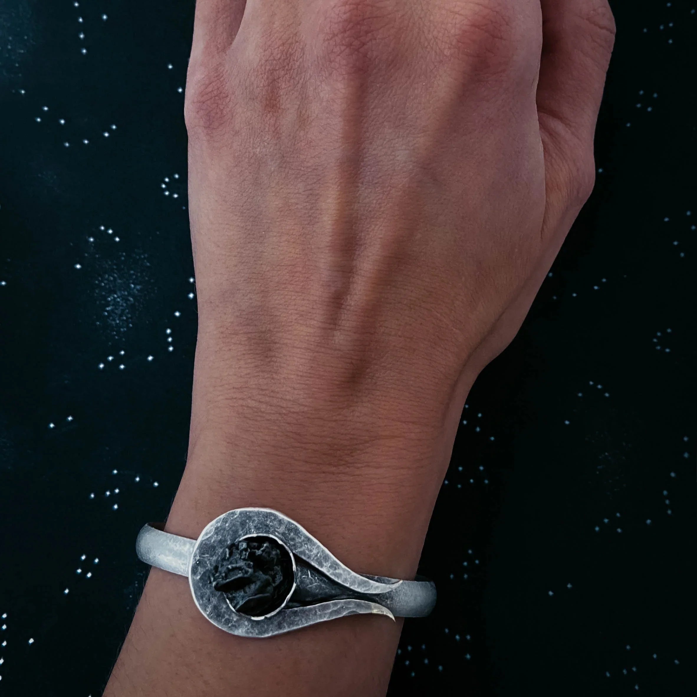 Close-up of a hand wearing a silver metal bracelet with a rustic black stone on a dark starry background