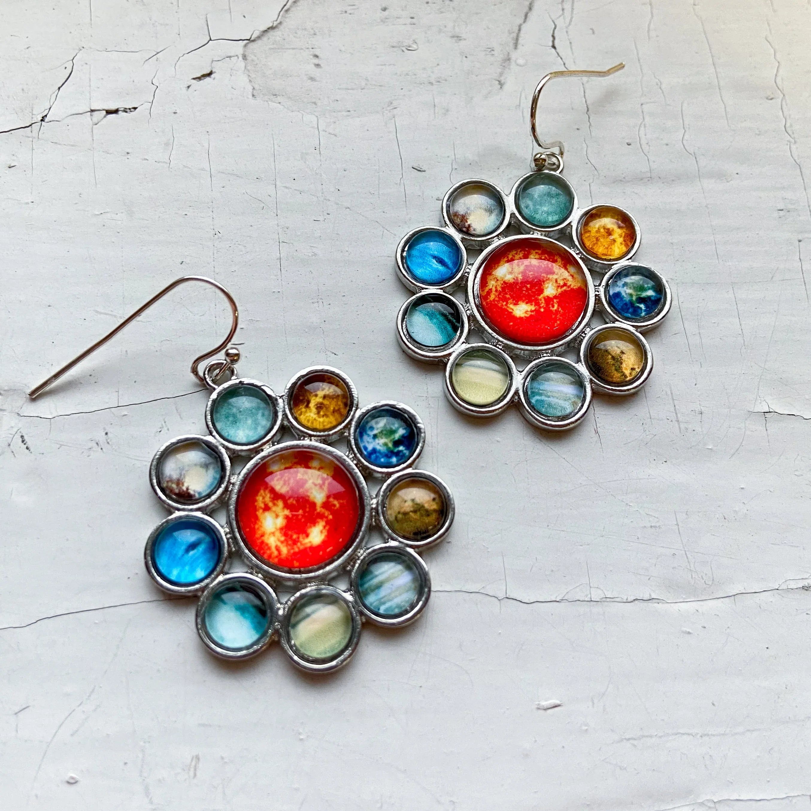 Silver earrings with multicolored planet designs arranged in a circular pattern on a cracked white surface