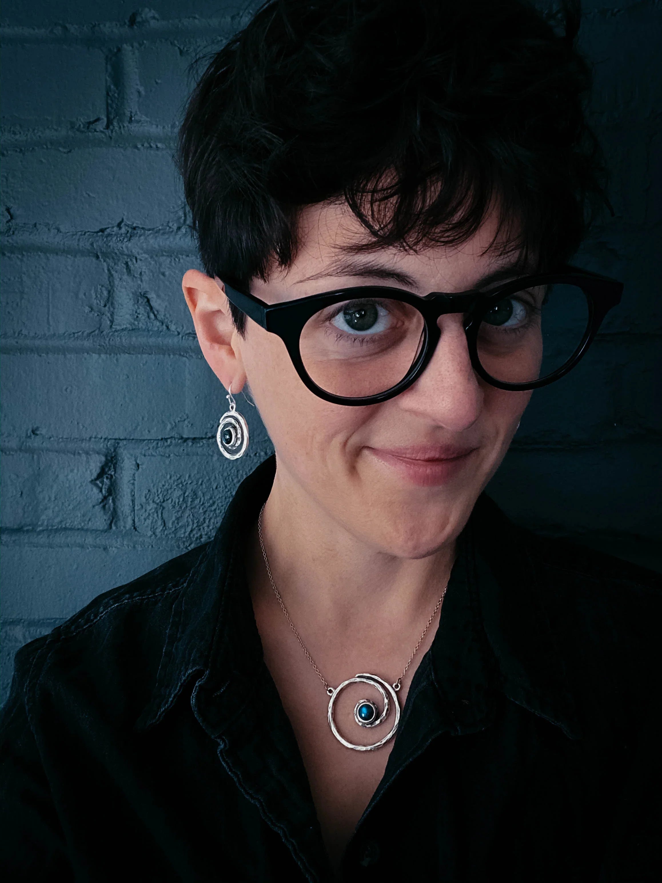 Woman in black glasses wearing cosmic-inspired silver necklace and earrings, dark brick background