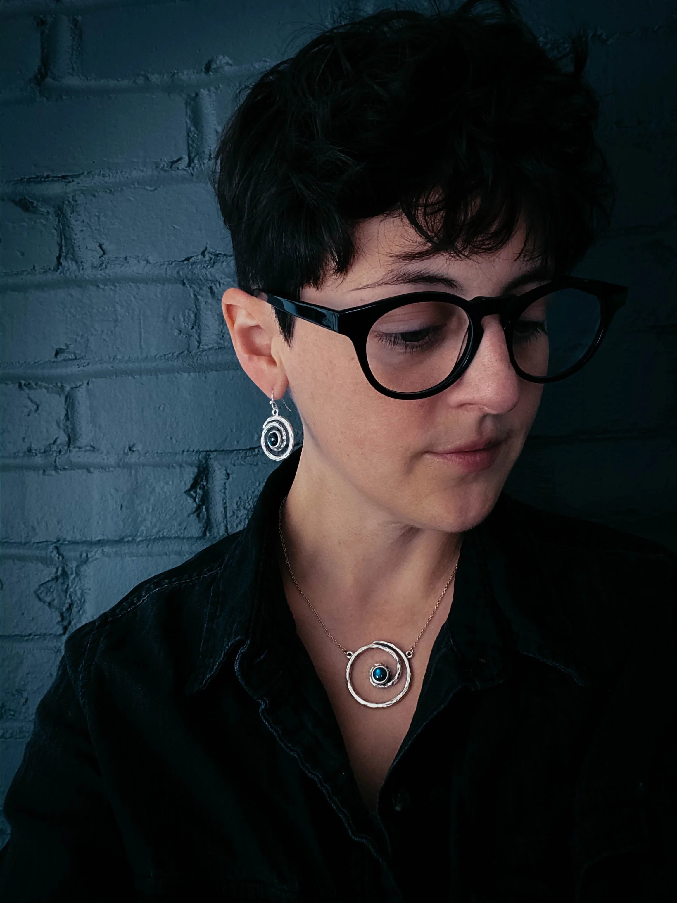 Woman with short dark hair wearing black glasses, cosmic spiral jewelry, and a dark shirt