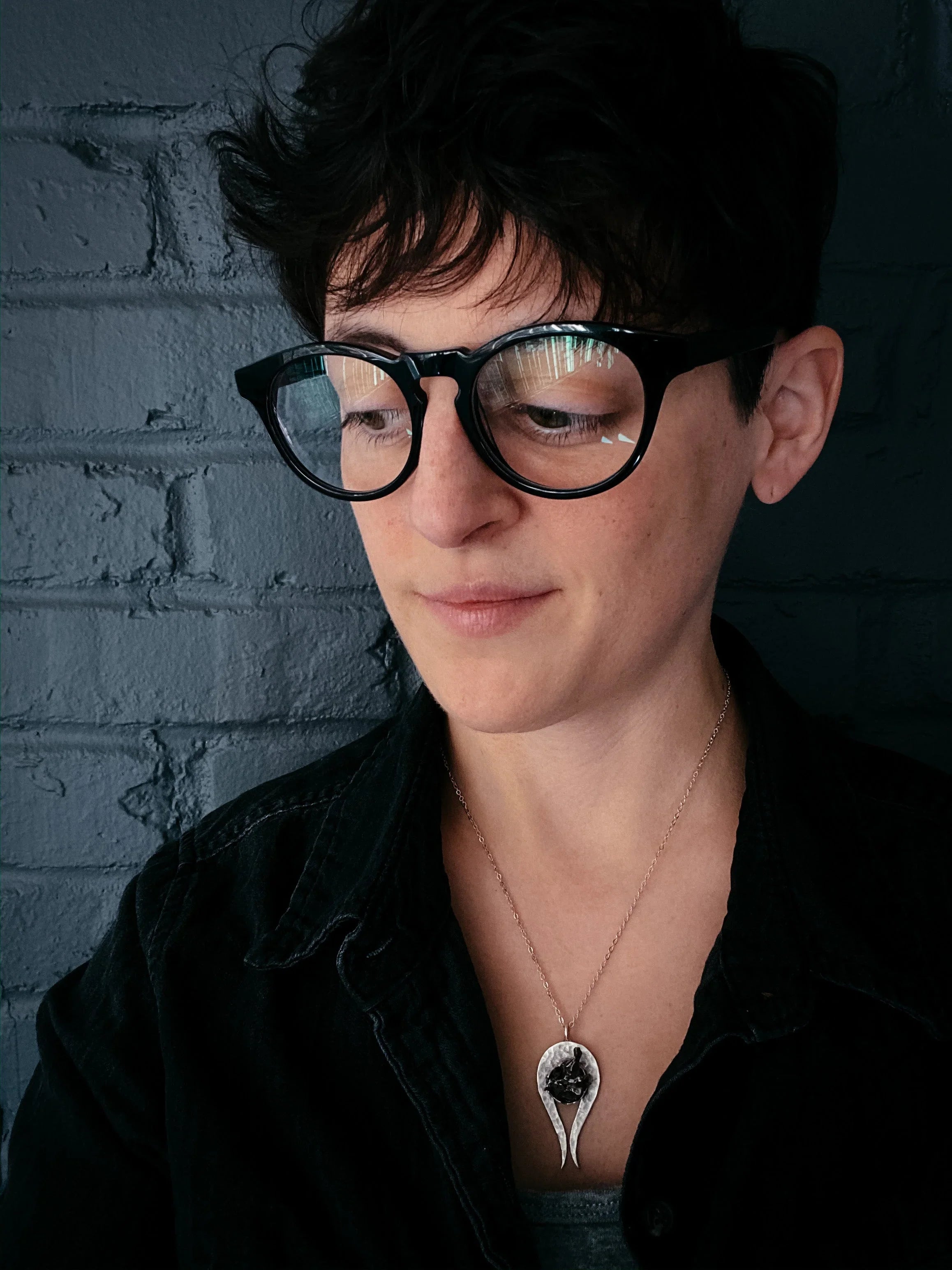 Person with short dark hair wearing black frame glasses and a unique pendant necklace against a dark brick wall
