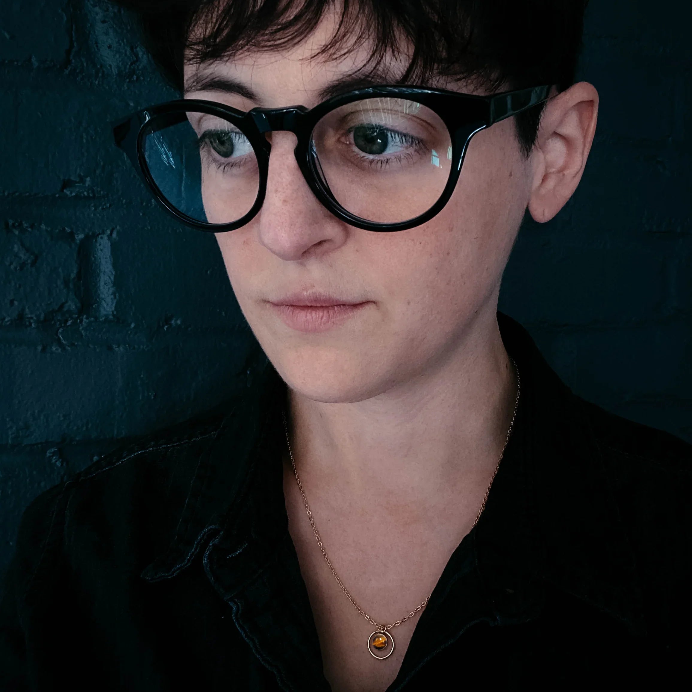Person with short hair, black glasses, and cosmic pendant necklace, against a dark brick wall