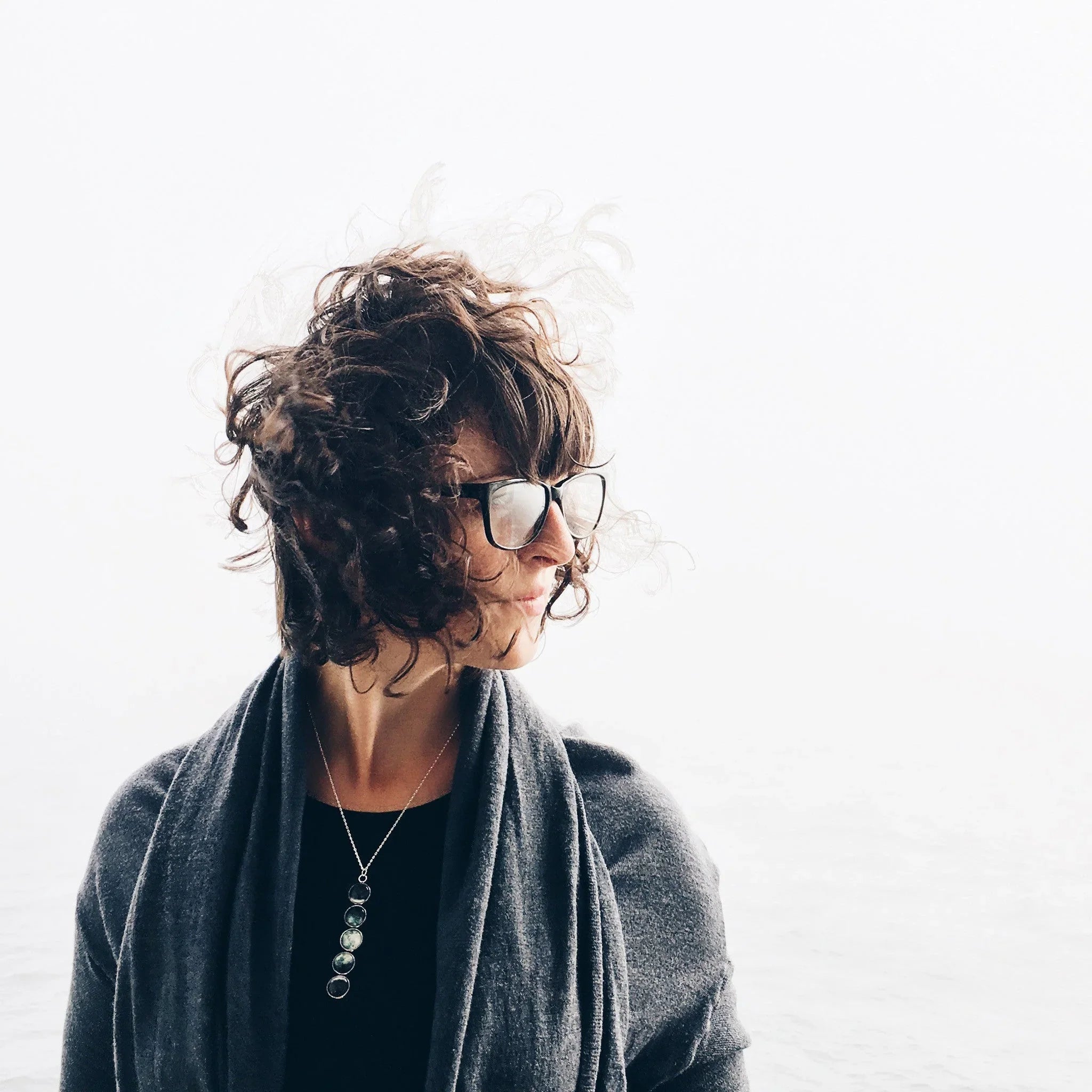 Woman with curly hair and glasses wearing dark clothing and pendant necklace against white background