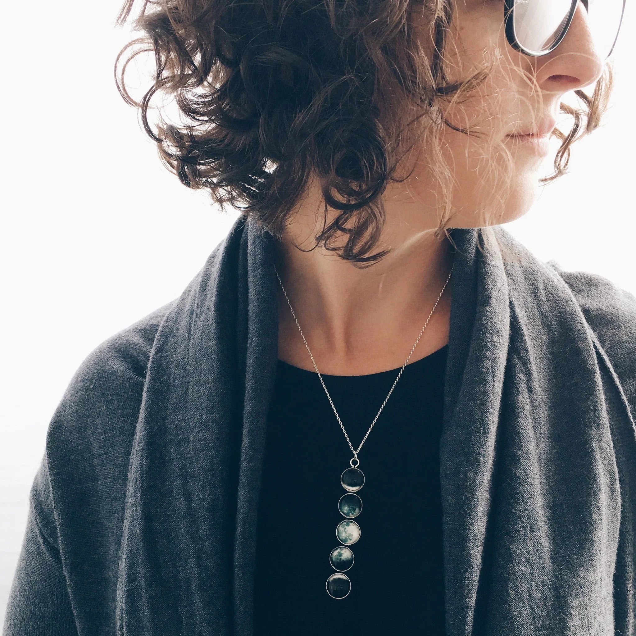 Woman with curly hair wearing round glasses, grey cardigan, and moon phase pendant necklace