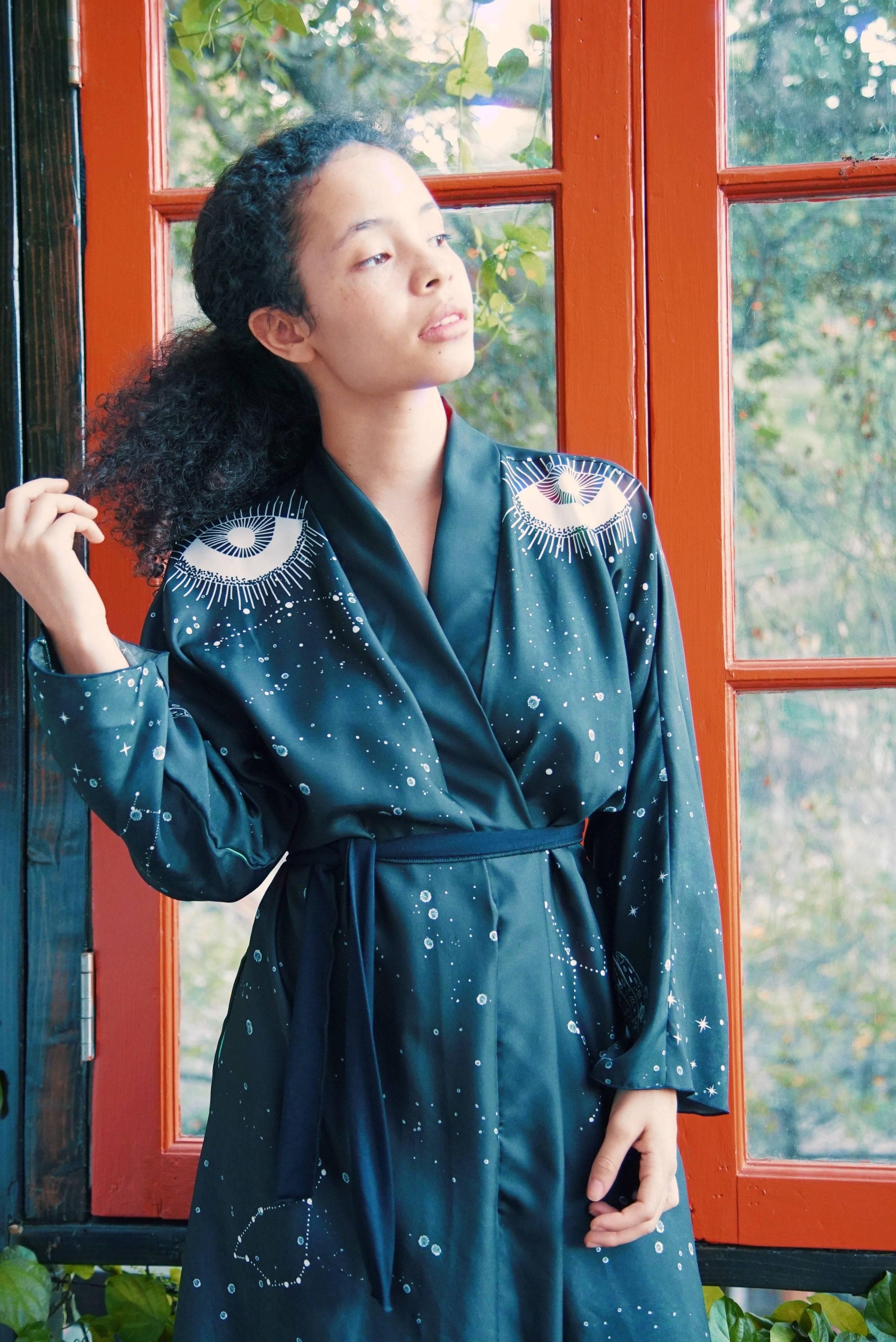 Woman in black celestial robe with eye designs posing by large orange-framed window with greenery outside