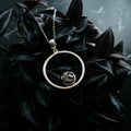Artist-designed silver pendant necklace with circular charm, displayed on dark foliage