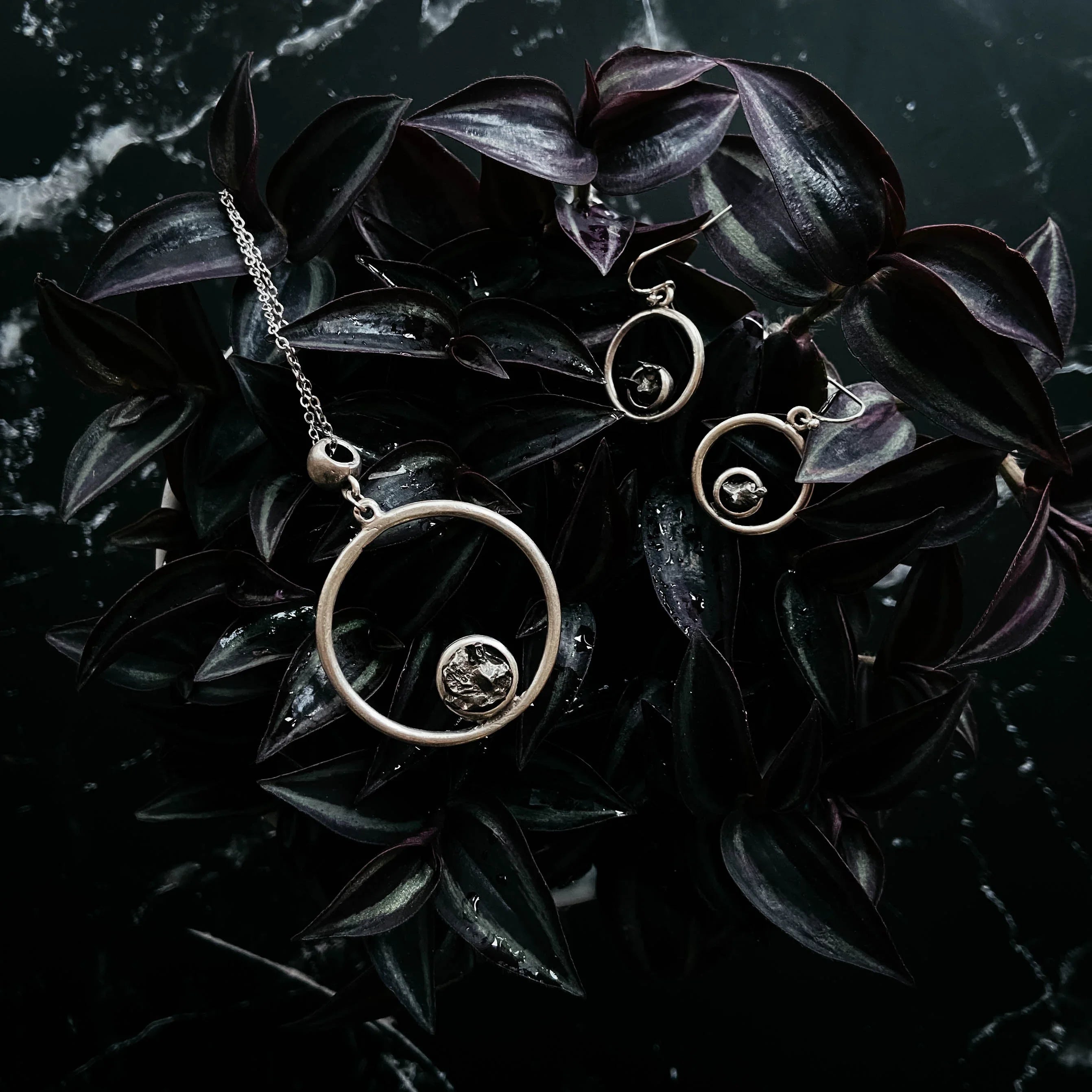 Silver circular pendant necklace and matching earrings on dark purple leaves with water droplets on marble surface