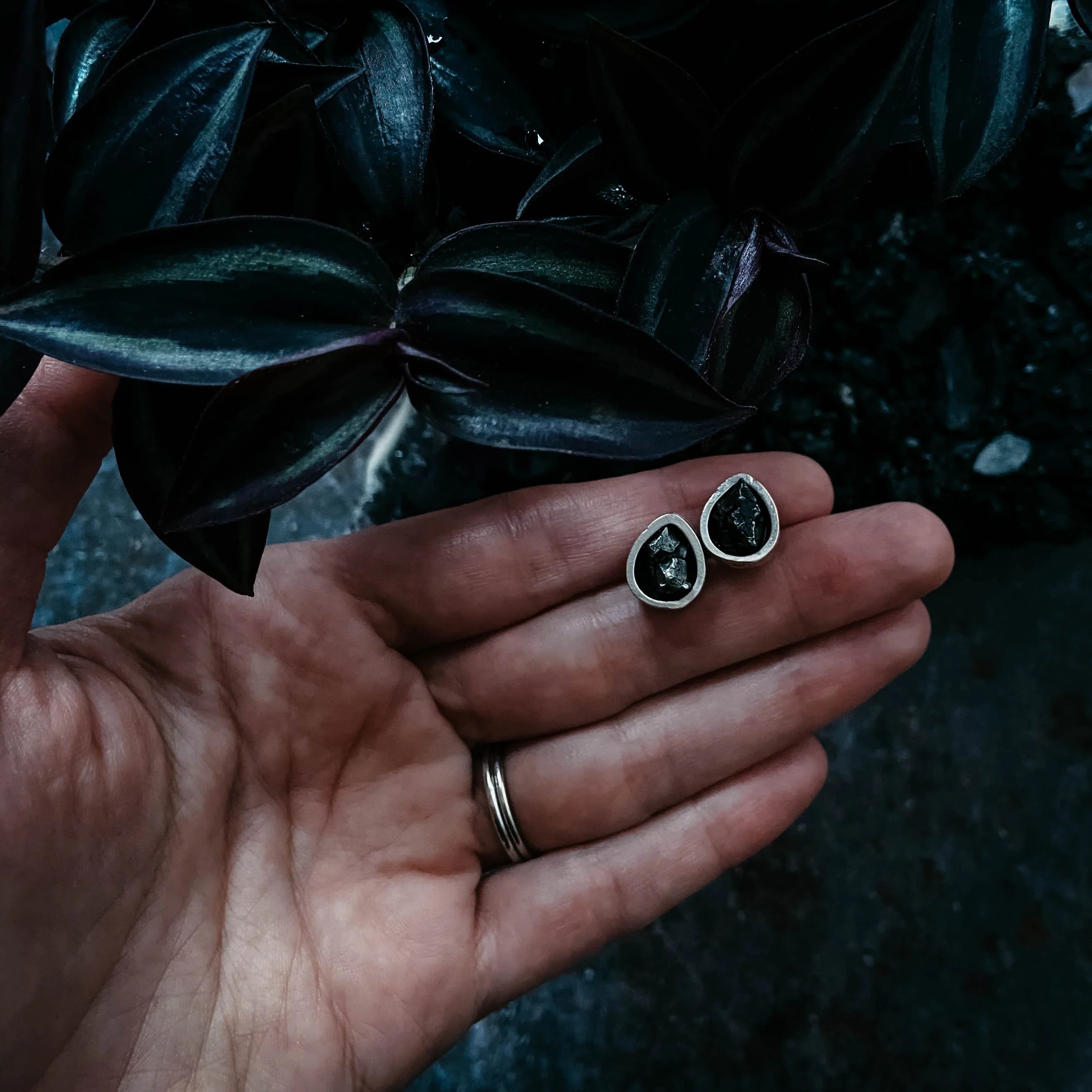 Hand holding teardrop black gemstone stud earrings near dark green plant at Qu!rk Boutique