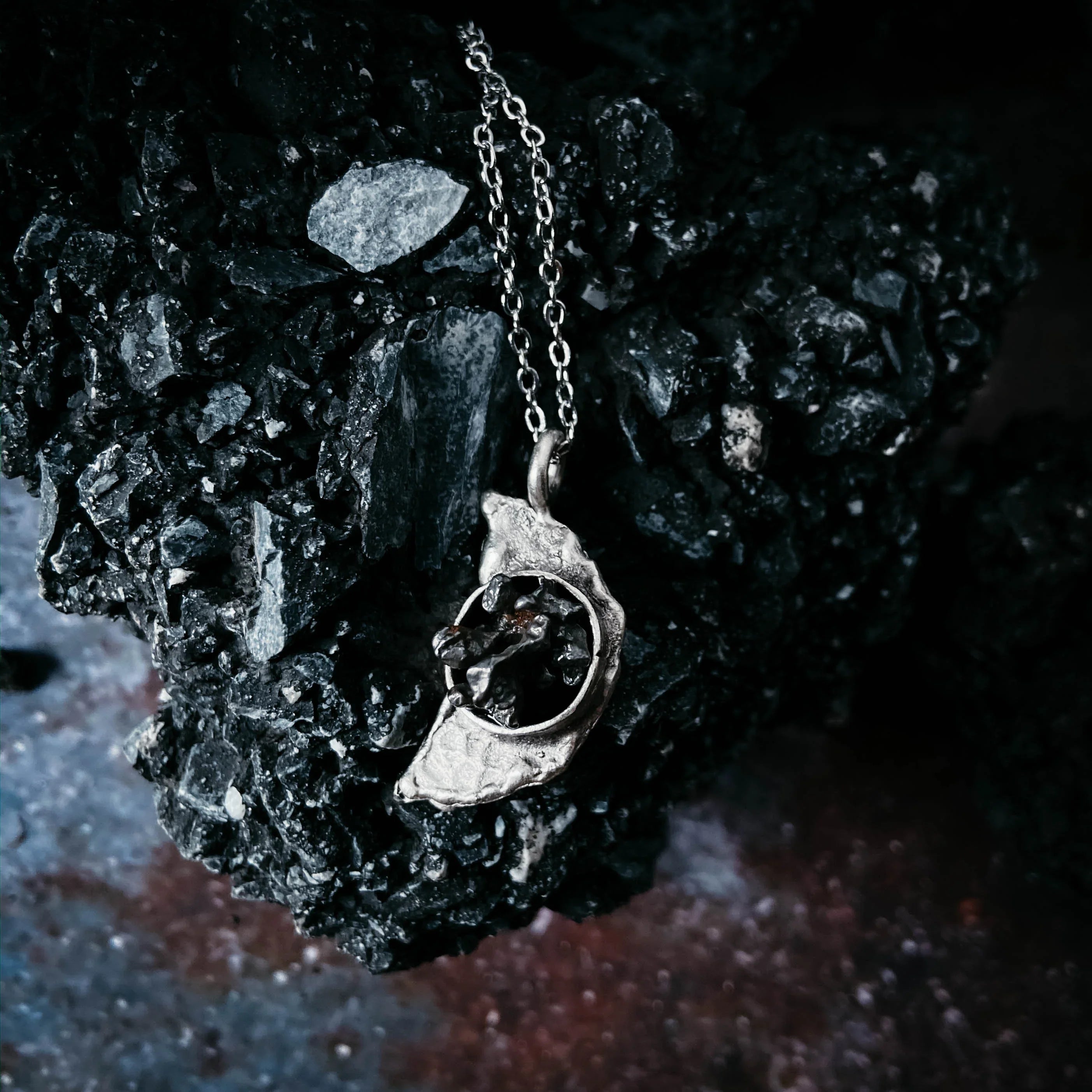 Silver pendant with black stone cluster on a silver chain resting on dark textured rock