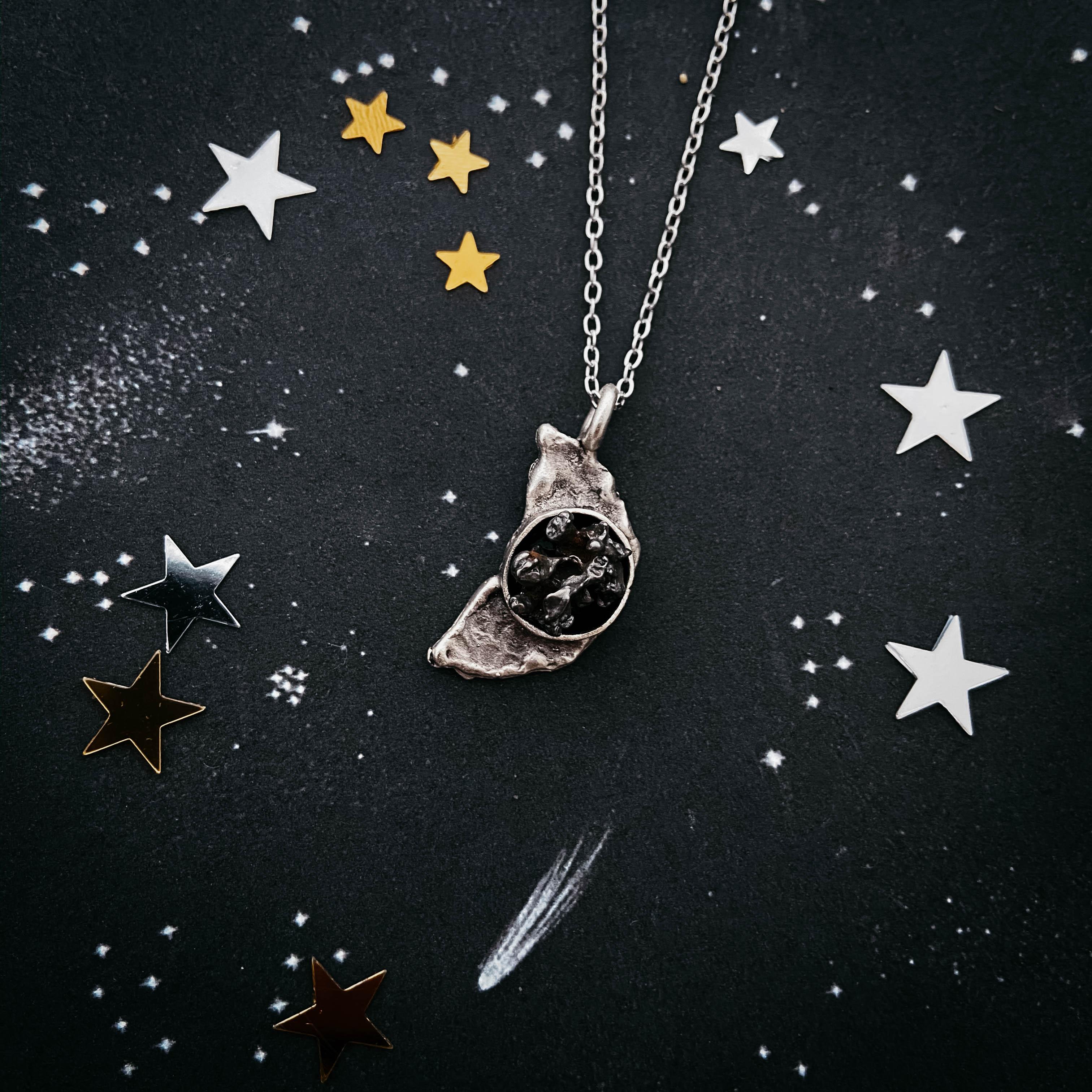 Silver moon-shaped pendant with black textured center on starry black background with metallic star decorations
