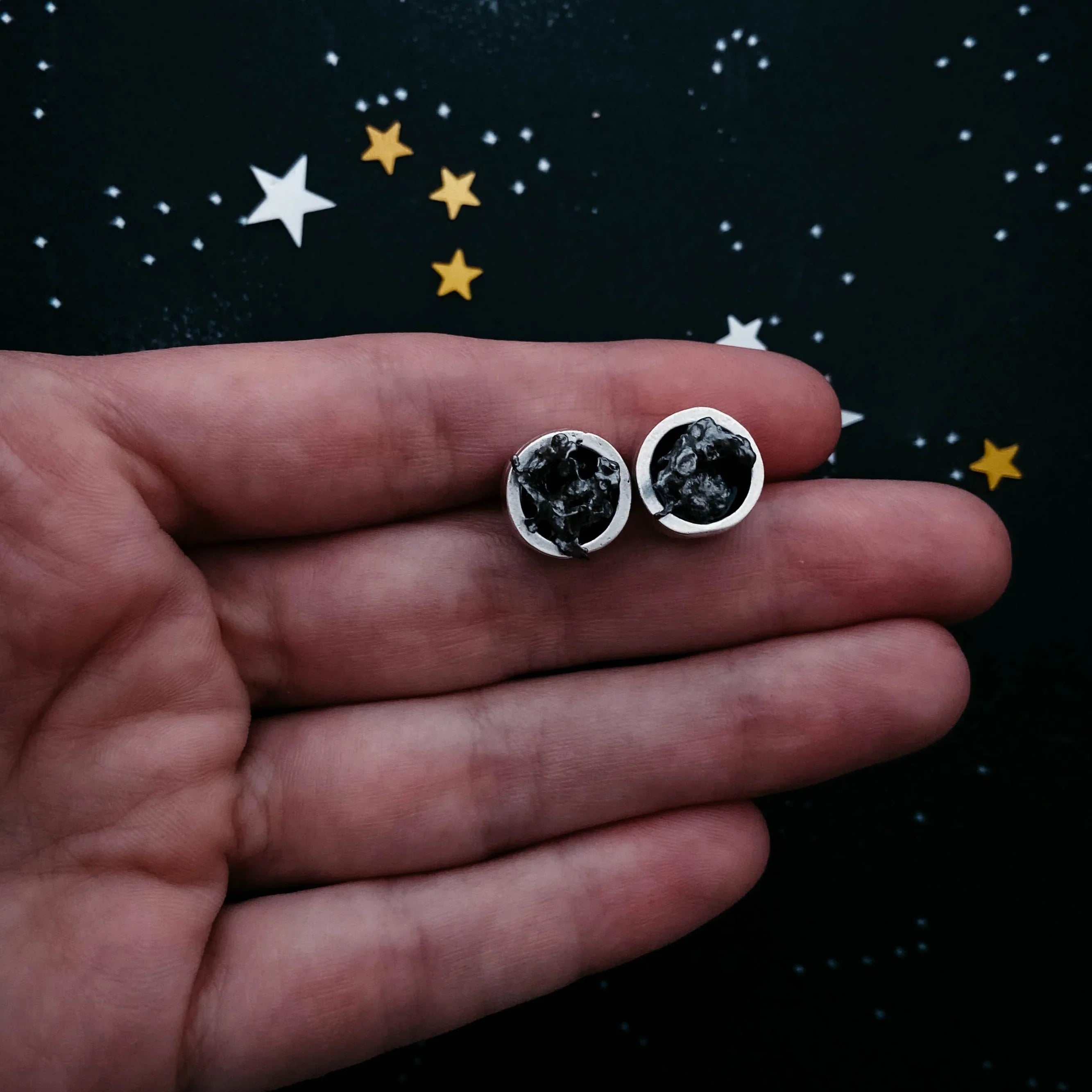 Hand holding a pair of silver stud earrings with black textured stones on starry background